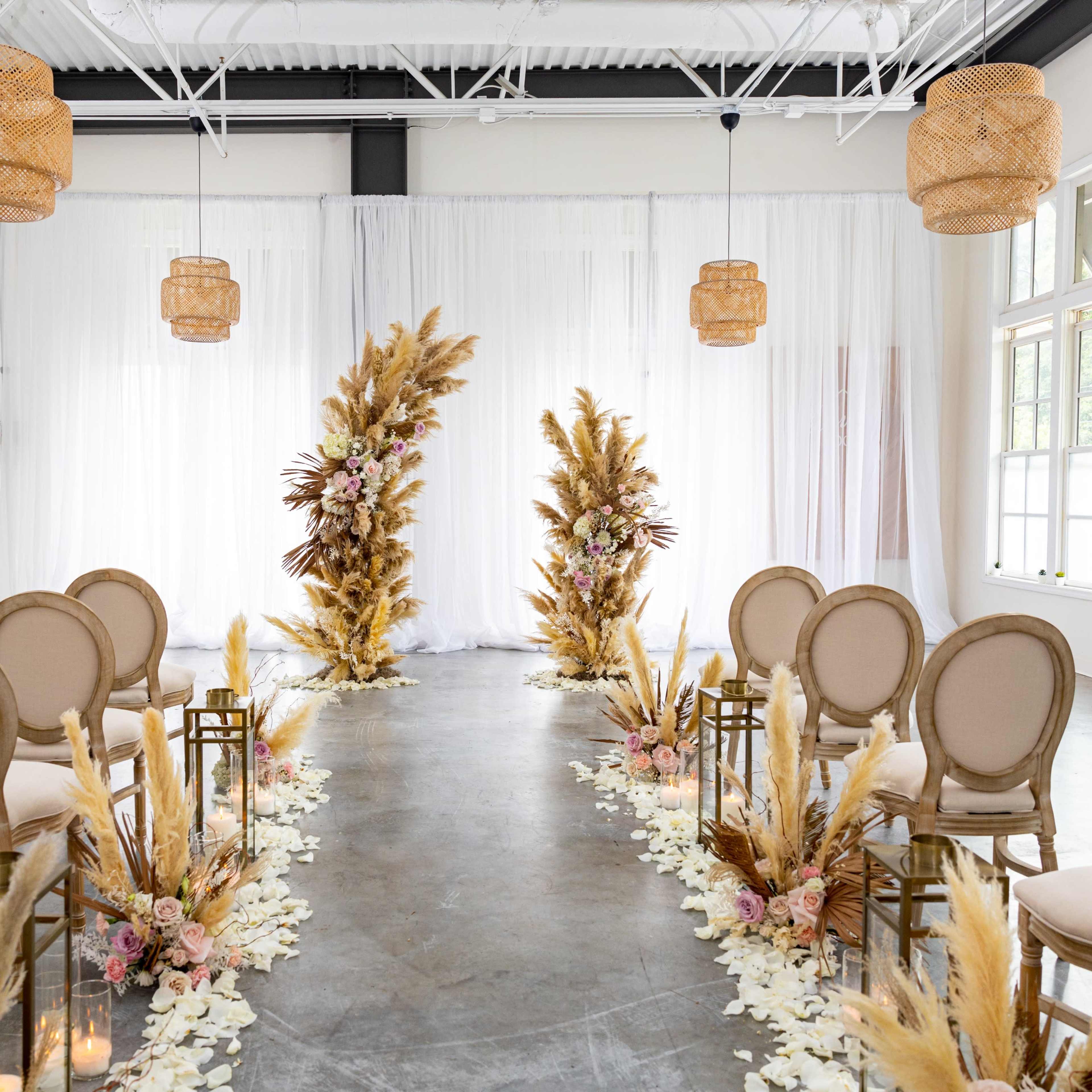 A wedding ceremony setup featuring rows of chairs flanking a floral arch made of pampas grass and other decorations, all set in a bright, white-walled venue.