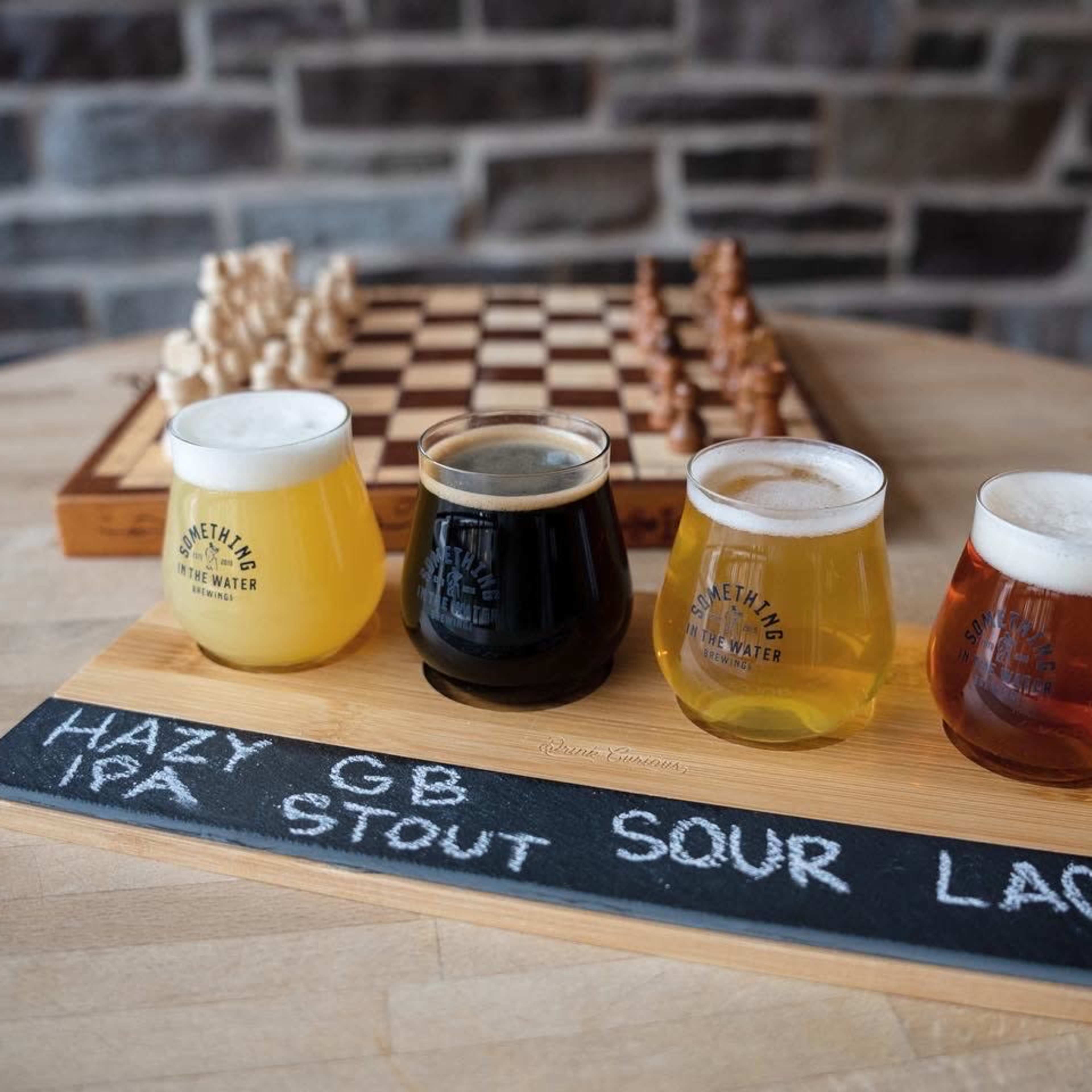 A wooden flight of four craft beers is displayed next to a chessboard on a stone wall background.