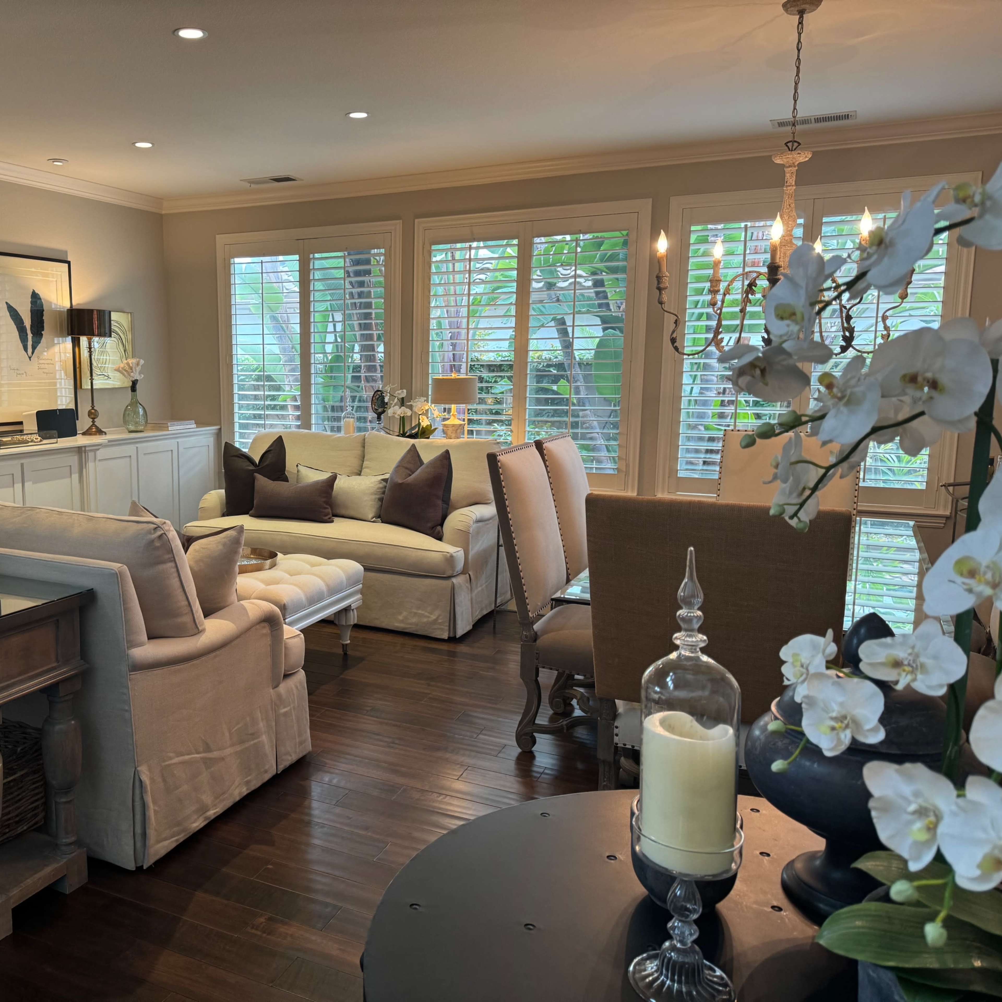 A well-furnished living room featuring a light-colored sofa, a wooden coffee table, and large windows letting in natural light, alongside decorative plants and artwork.