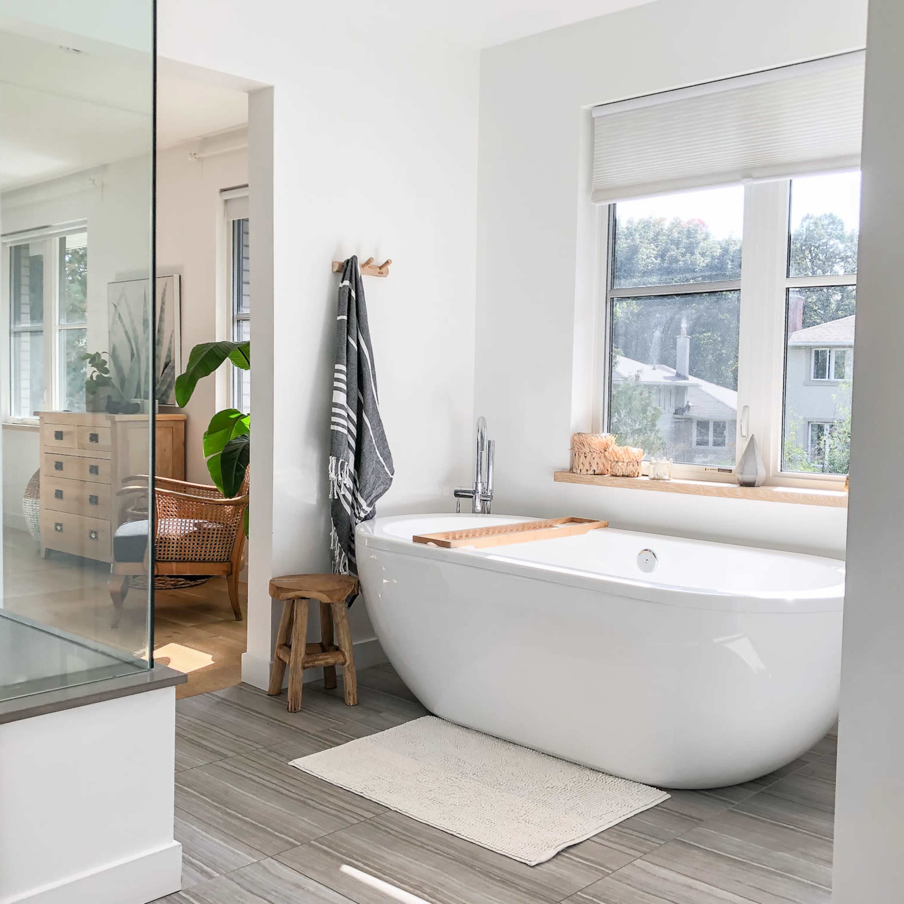 A modern bathroom featuring a freestanding white bathtub, a wooden stool, a large window with natural light, and minimal decor.
