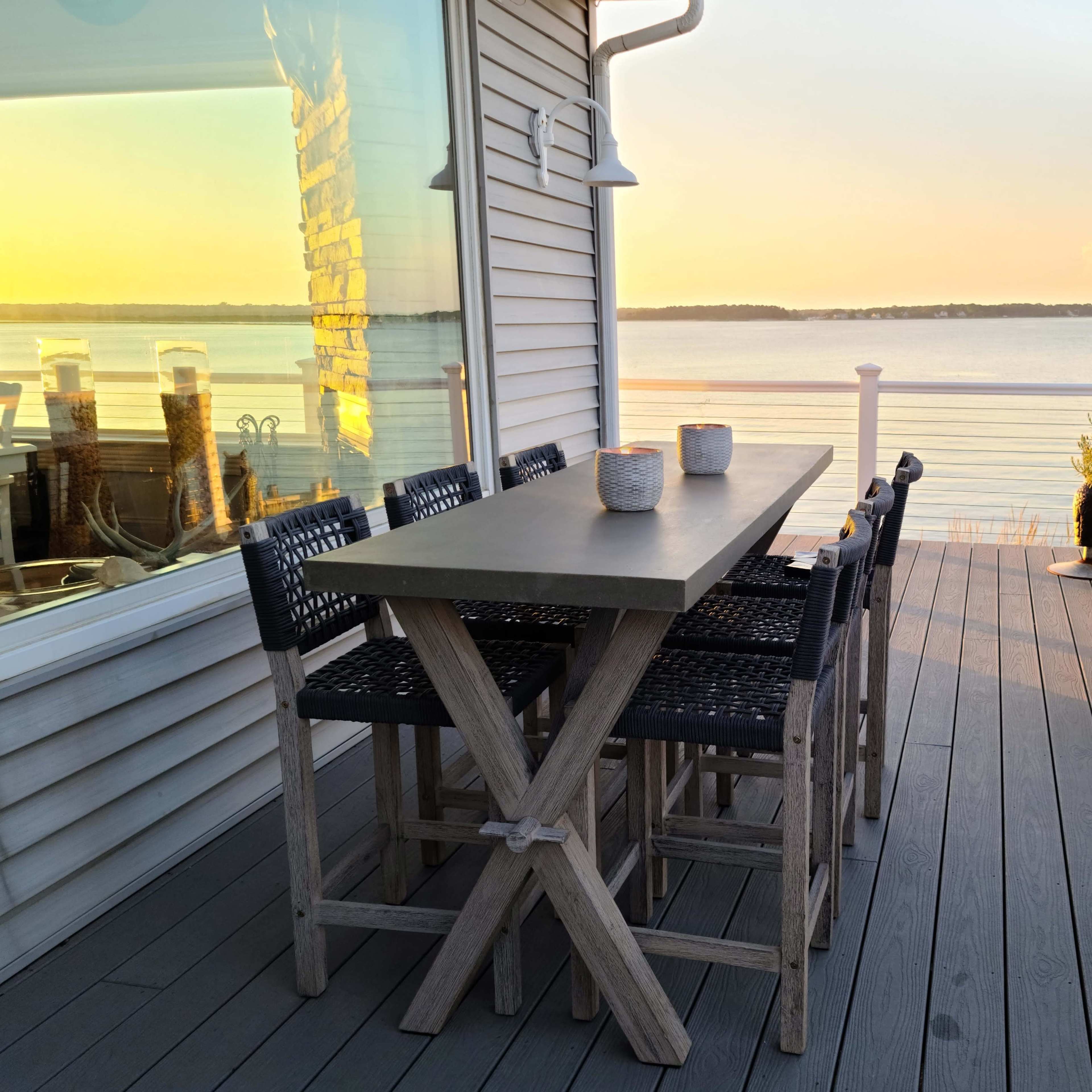 A wooden table with black woven chairs stands on a deck overlooking a calm body of water at sunset.