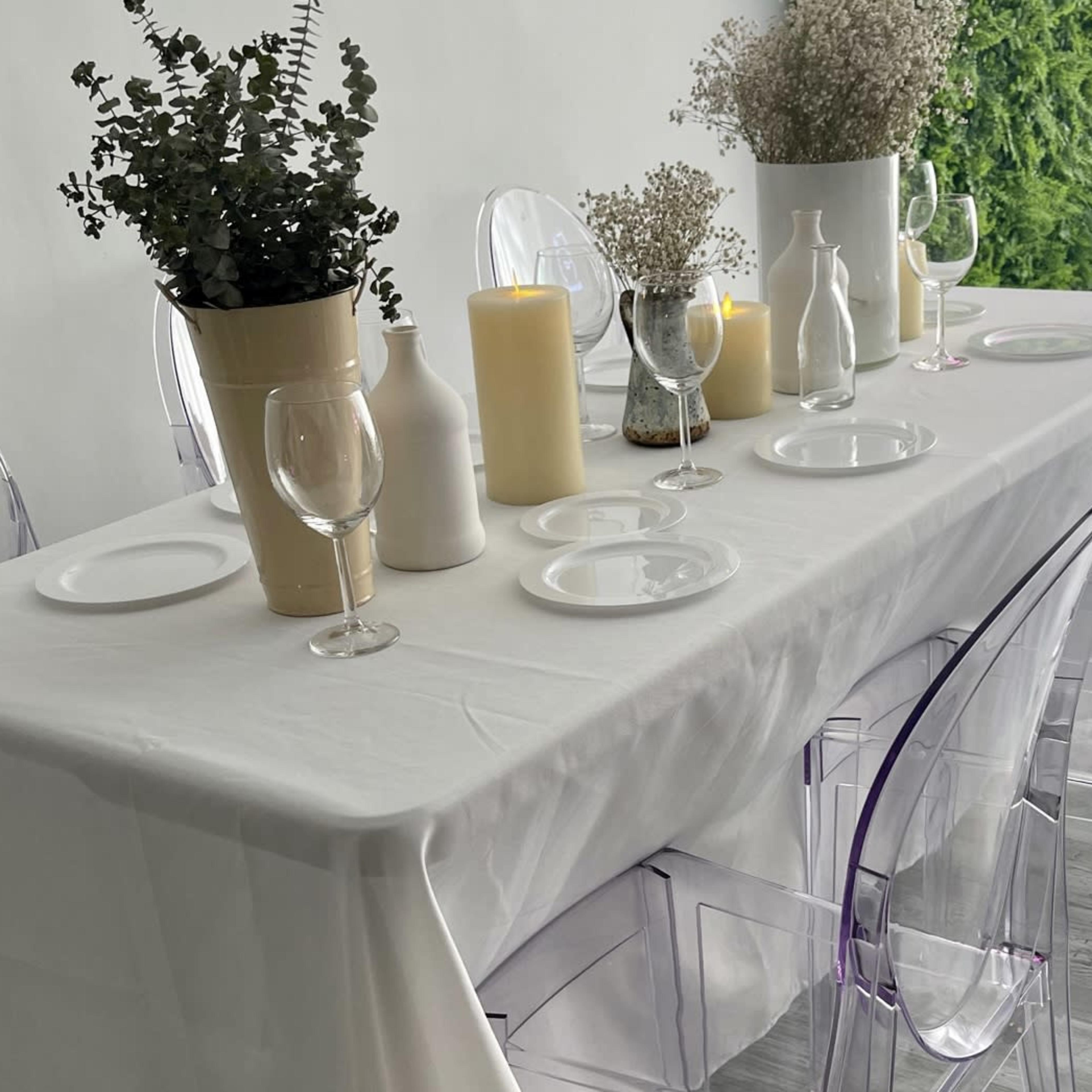 A long, white table with clear chairs is set in a bright room featuring a green plant wall and neutral decor.