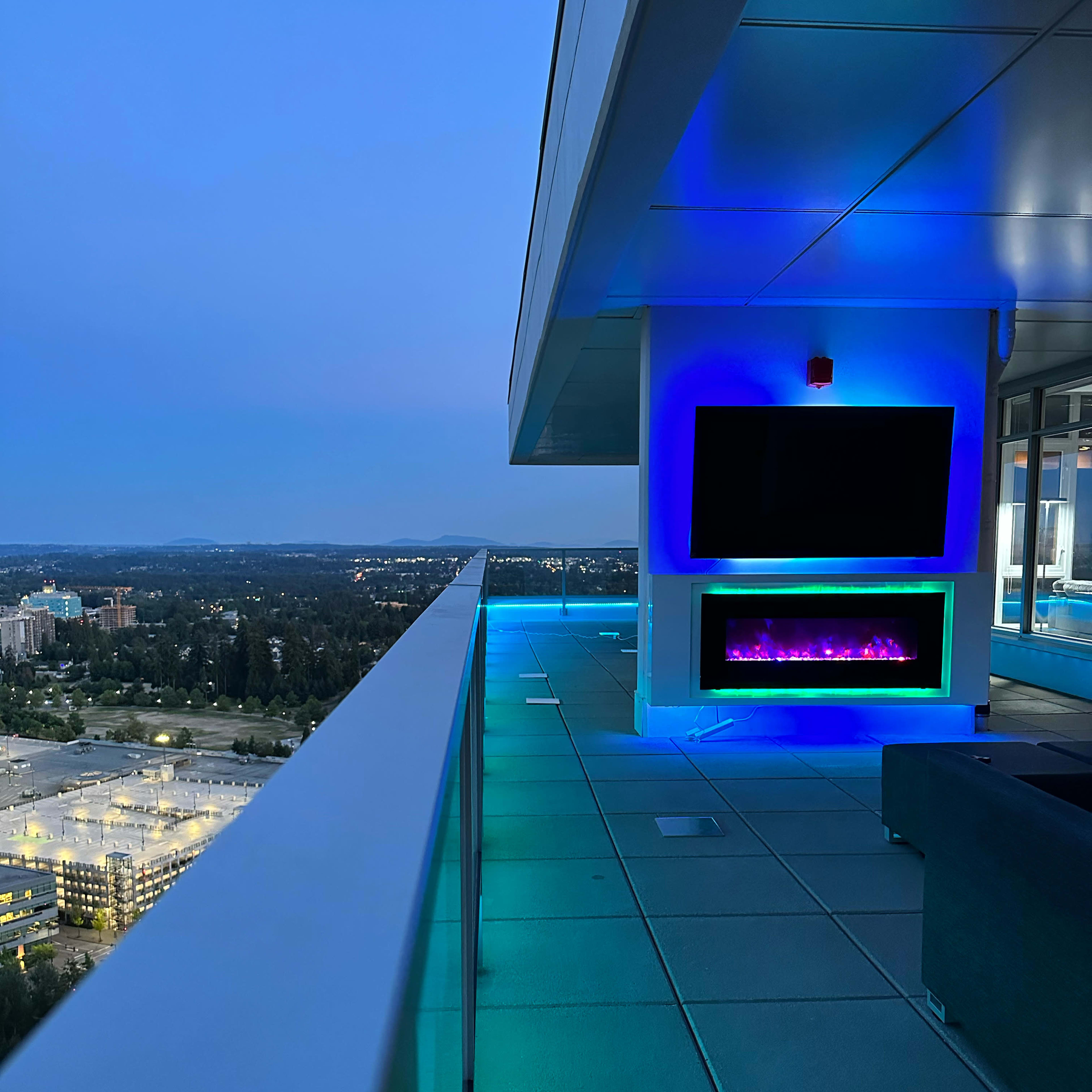 A modern balcony with a wall-mounted TV and a fireplace, illuminated by blue and green lights, overlooks a cityscape at dusk.