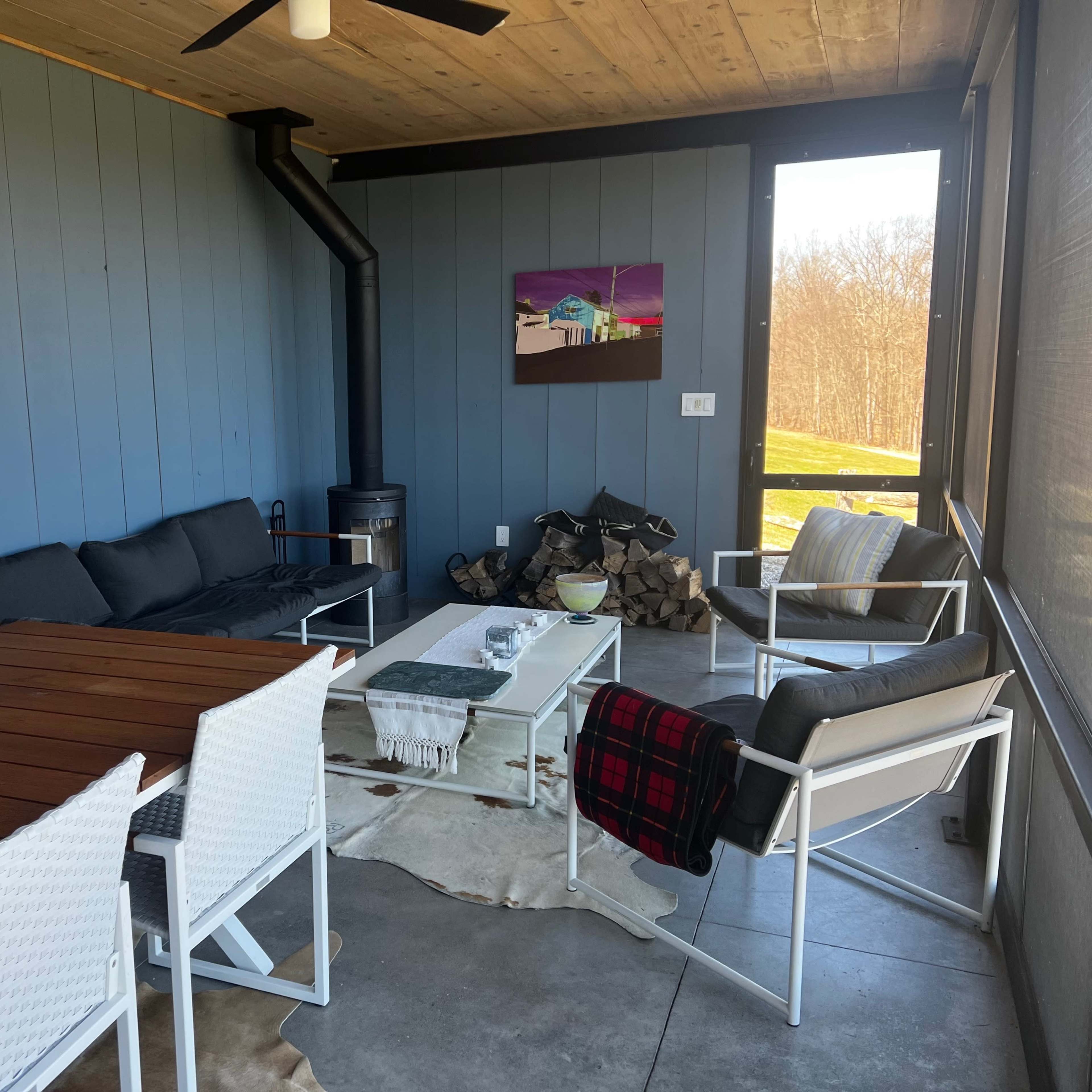 A covered patio features a dining table, seating area with a sectional sofa and accent chairs, a wood stove, and a decorative wall art piece.