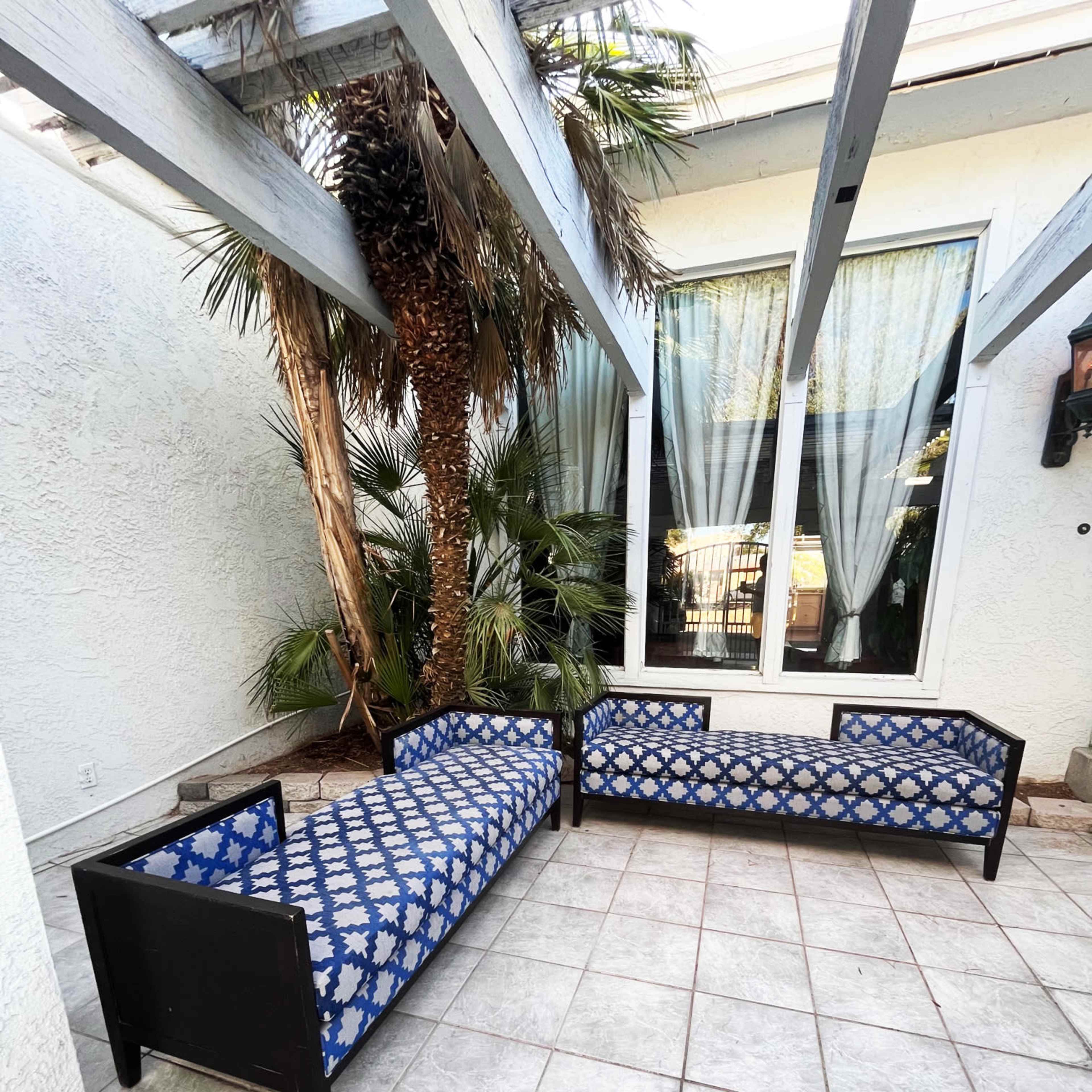 Two black couches with blue patterned upholstery set on a tiled patio surrounded by greenery and large windows.