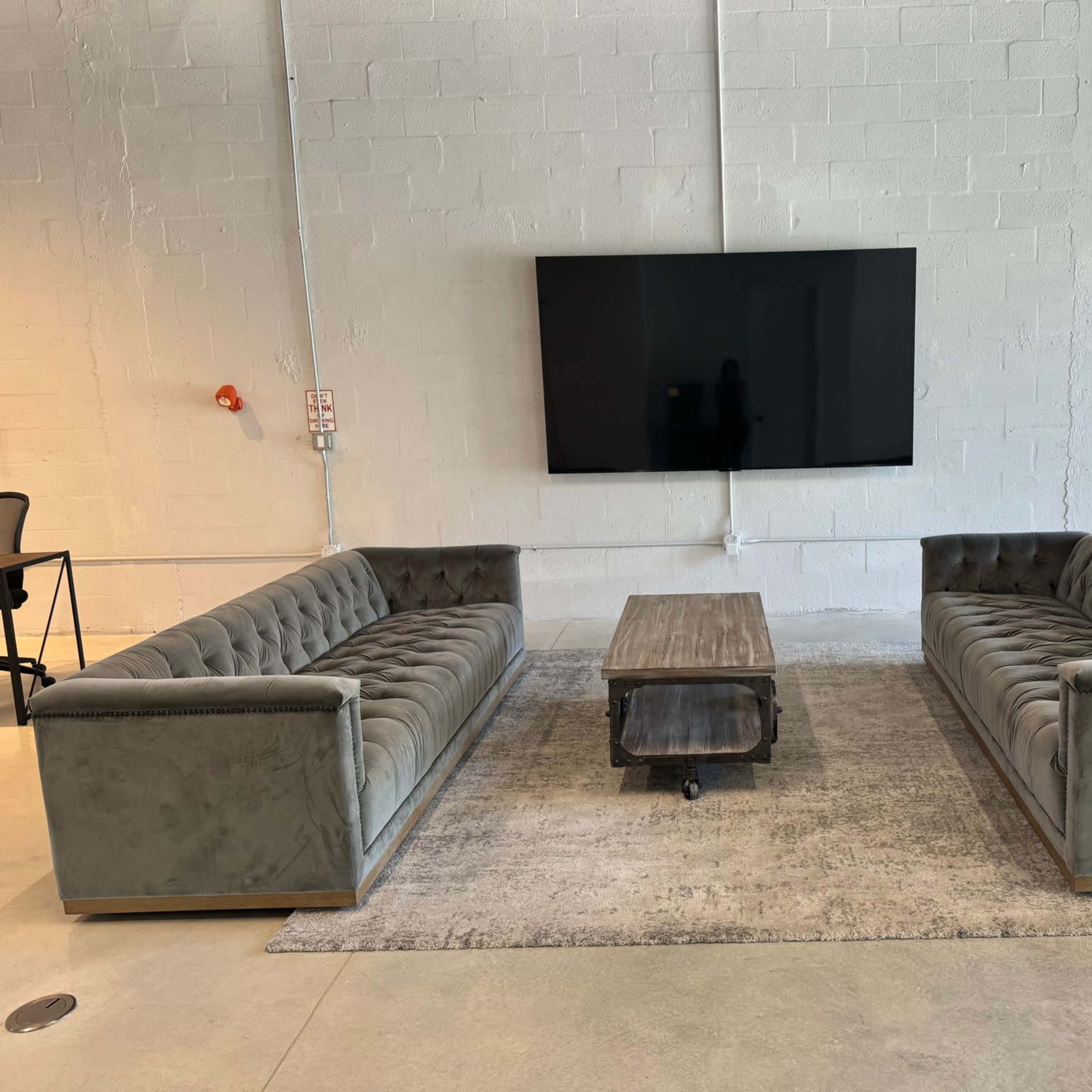 A furnished room with two tufted gray sofas facing a coffee table on a light rug, and a large flat-screen television mounted on a white wall.