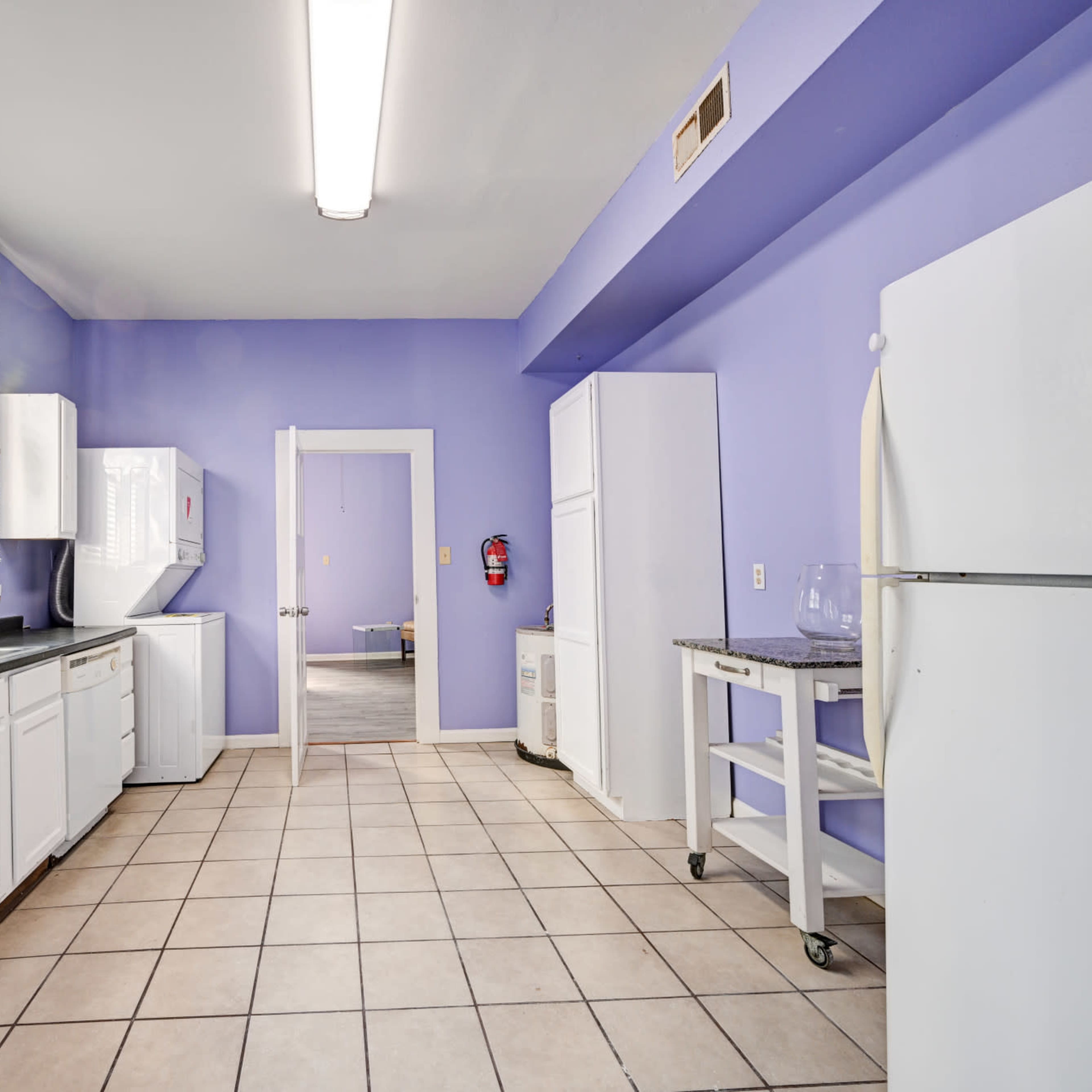 A kitchen with purple walls, white cabinetry, and tiled flooring, featuring a window with blinds, a washer and dryer, and stainless steel and white appliances.