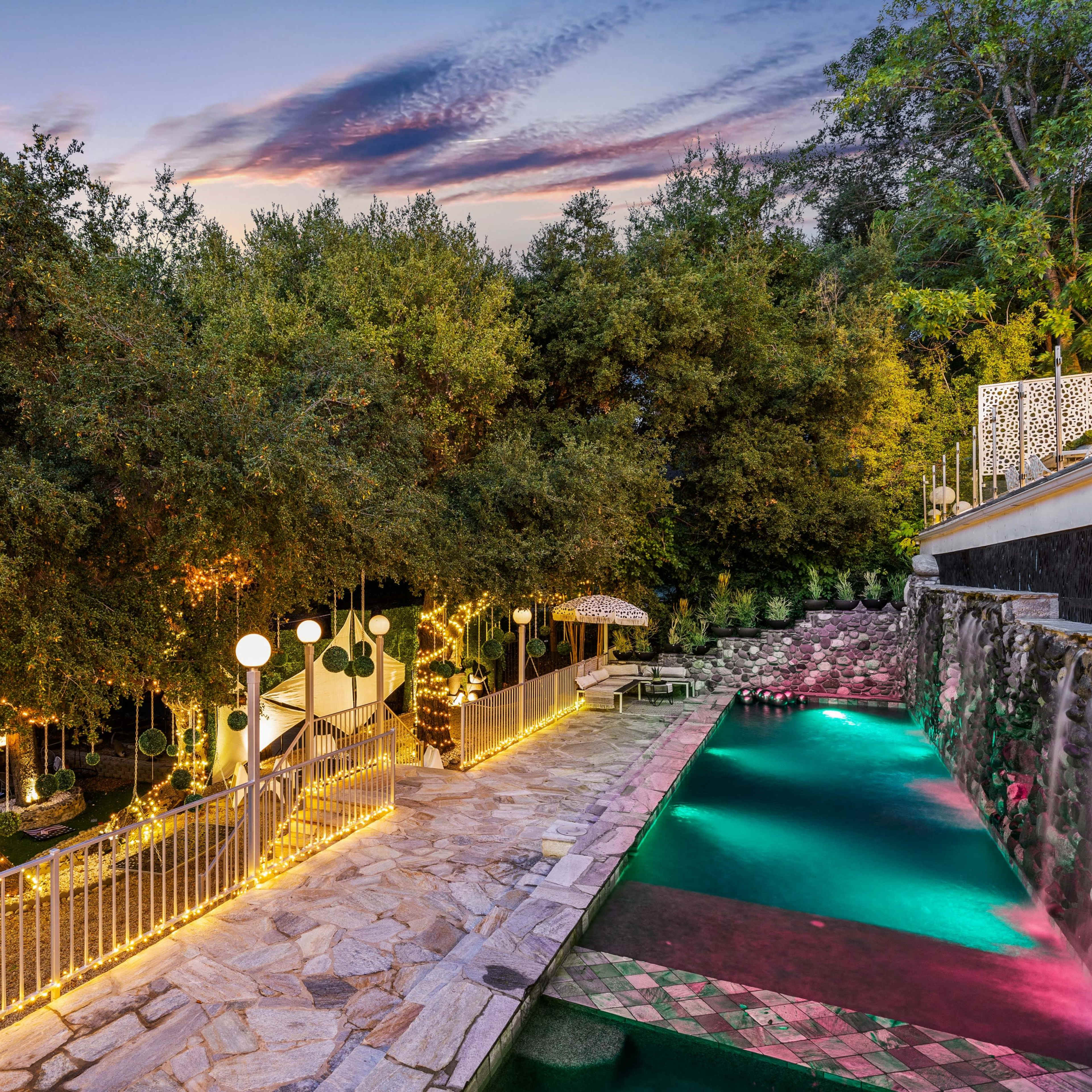 A landscaped backyard featuring a swimming pool, stone pathways, and decorative lighting along lush greenery at twilight.