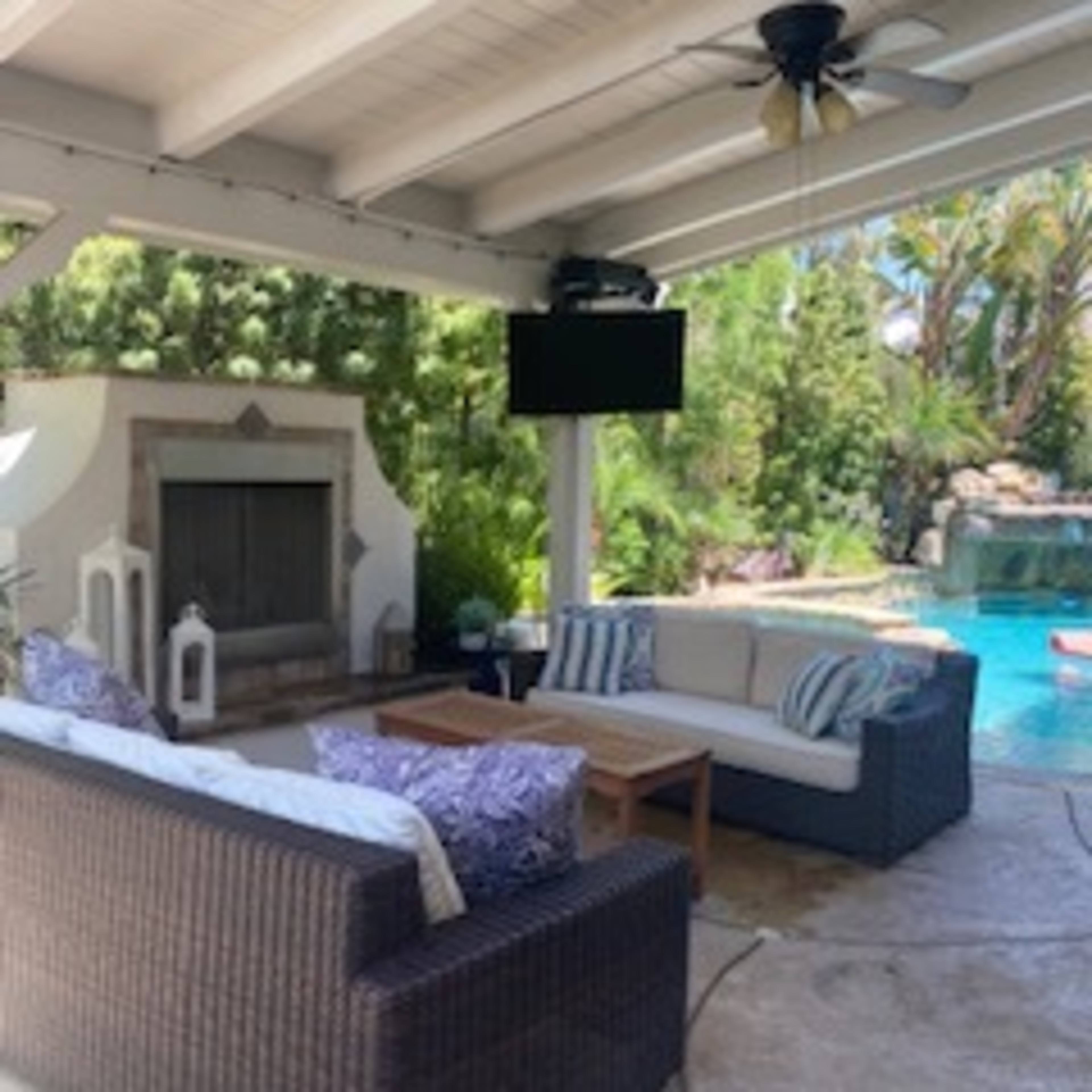 A covered patio features a seating area with couches and a coffee table, alongside a pool with lush greenery in the background.