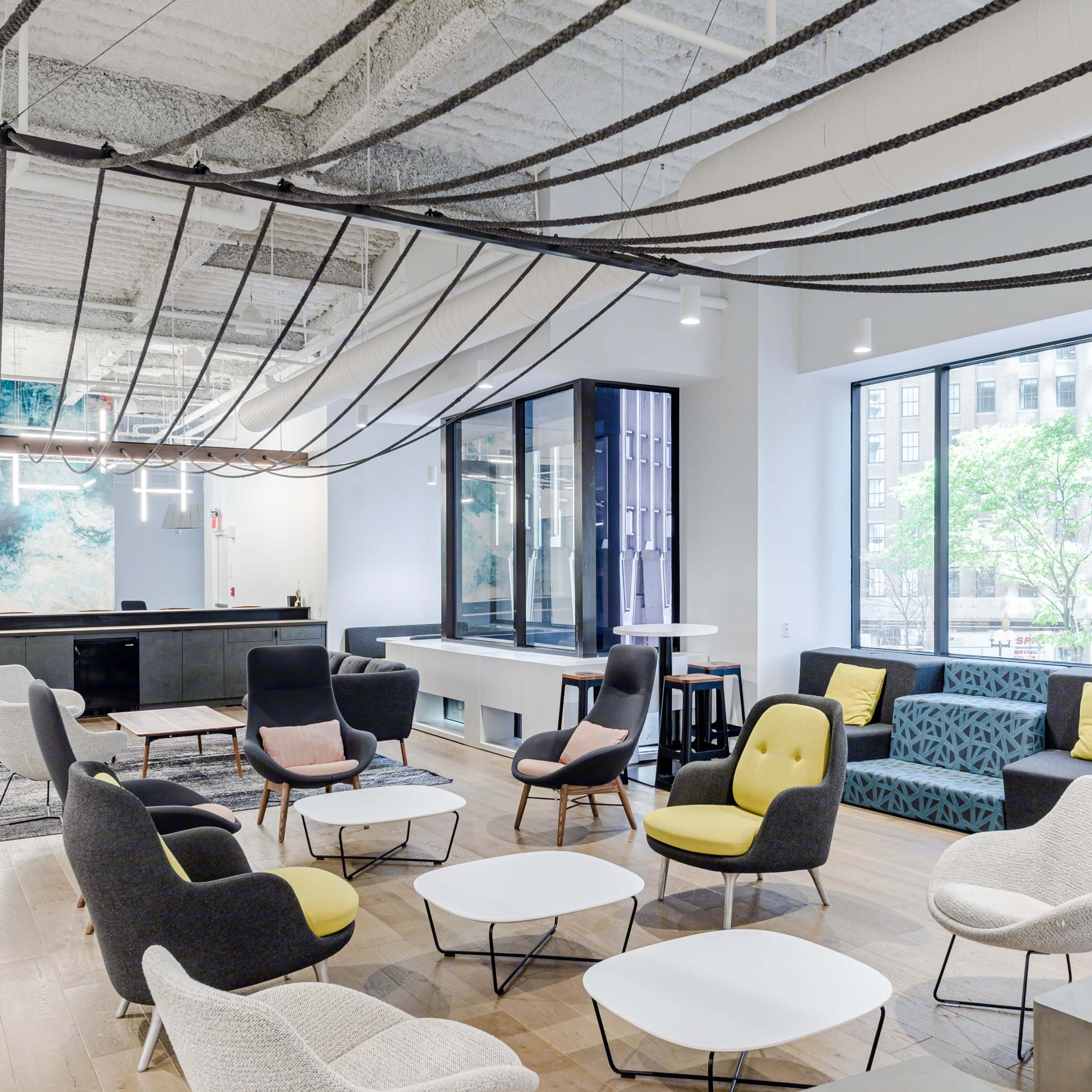 A modern office lounge area with various seating options, large windows, and suspended decorative elements above.