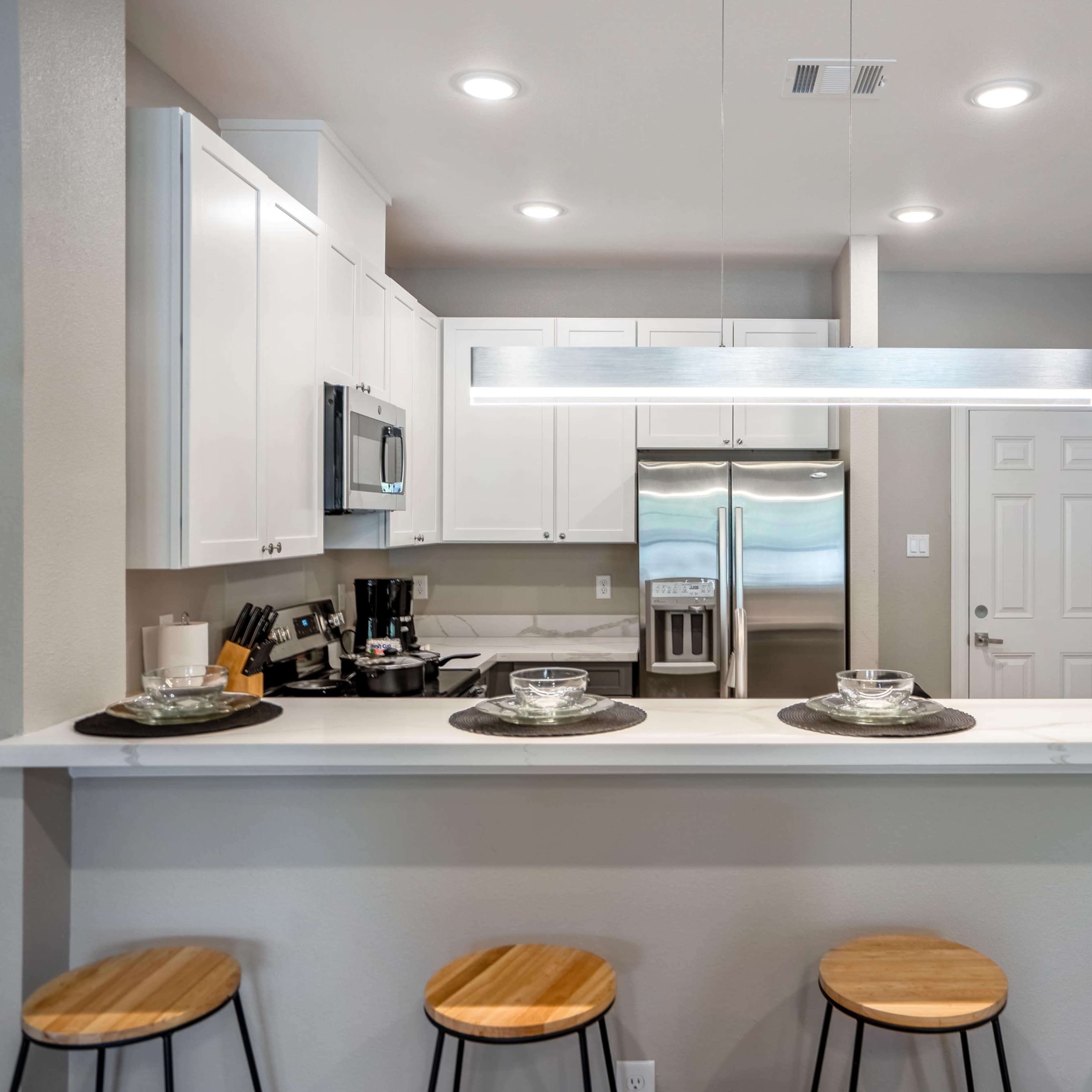 A modern kitchen features a counter with four wooden stools, sleek white cabinetry, and stainless steel appliances.