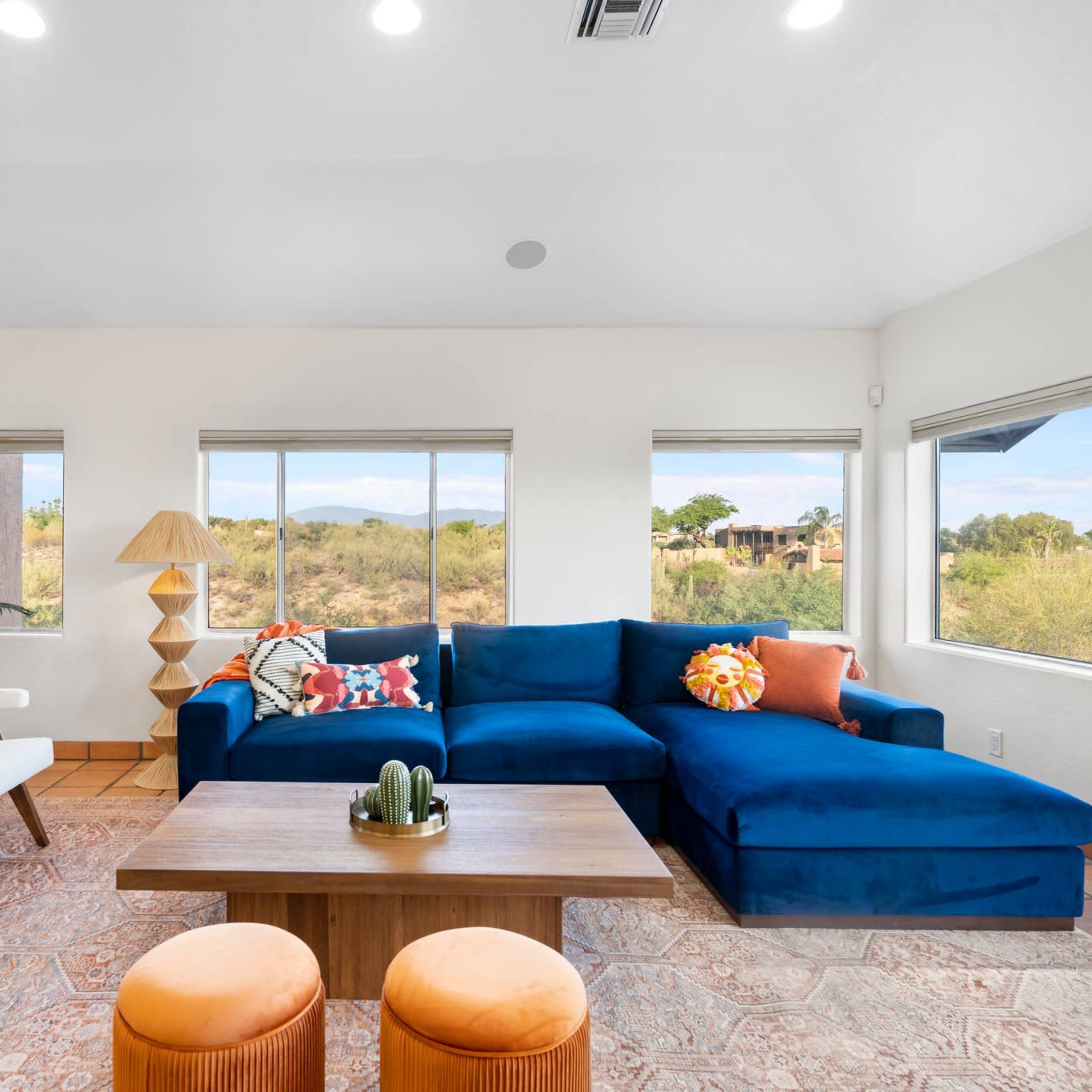 The living room features a blue sectional sofa, a wooden coffee table, and large windows that provide a view of the surrounding landscape.