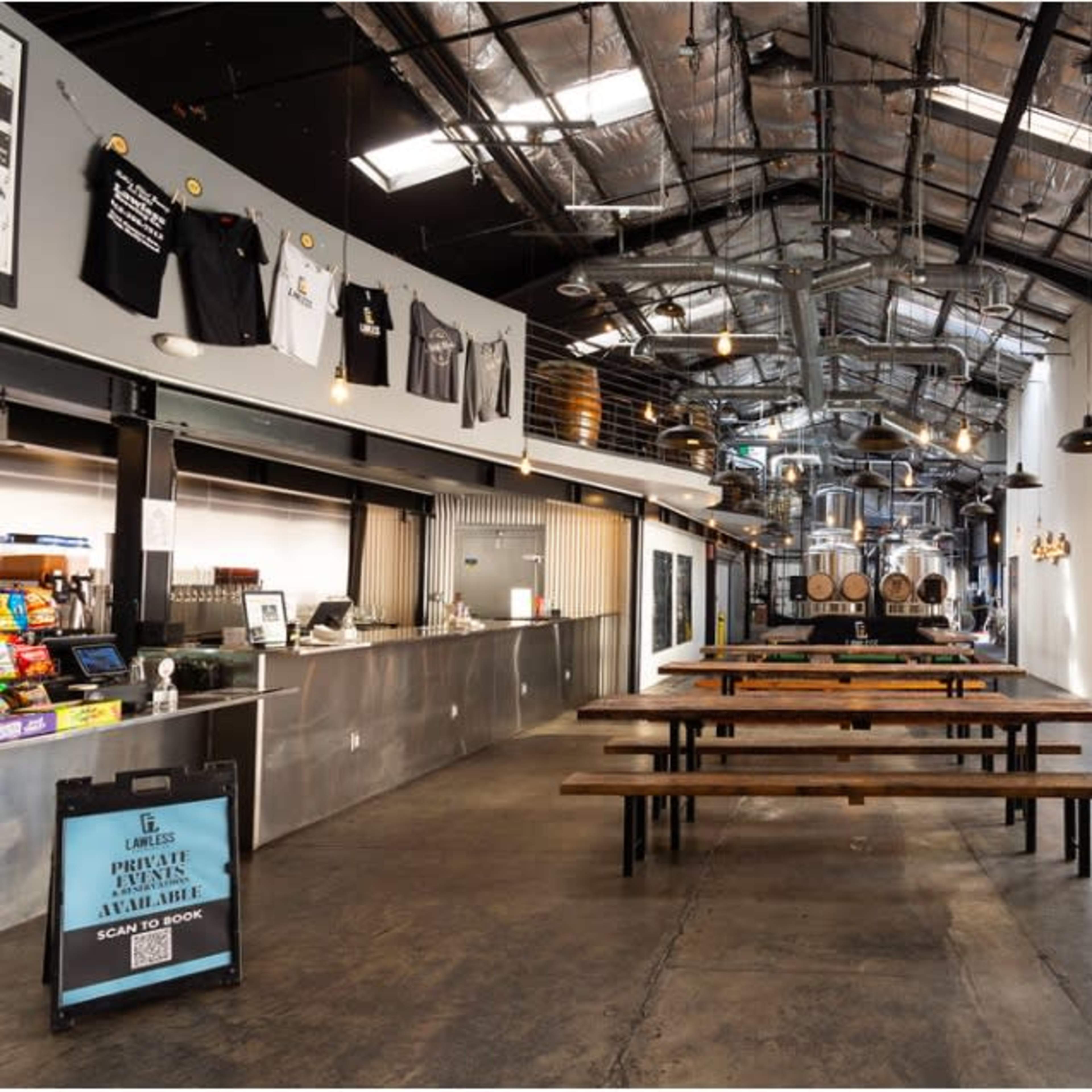 A spacious industrial-style dining area with long wooden tables, a counter with food service, and large windows that open to an outdoor area.