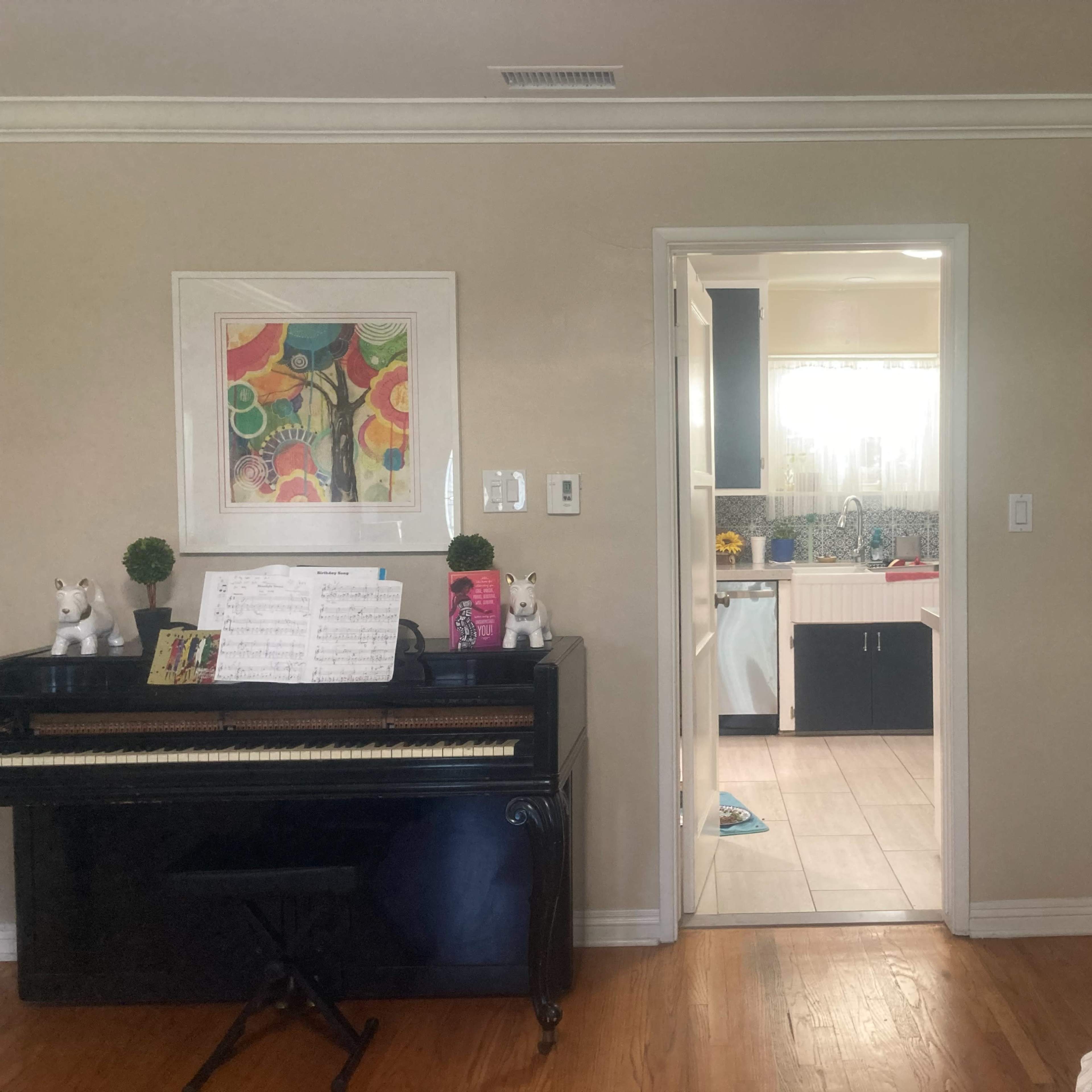 A black piano sits against a wall, with a doorway leading to a kitchen area visible in the background, and a plant in the corner of the room.