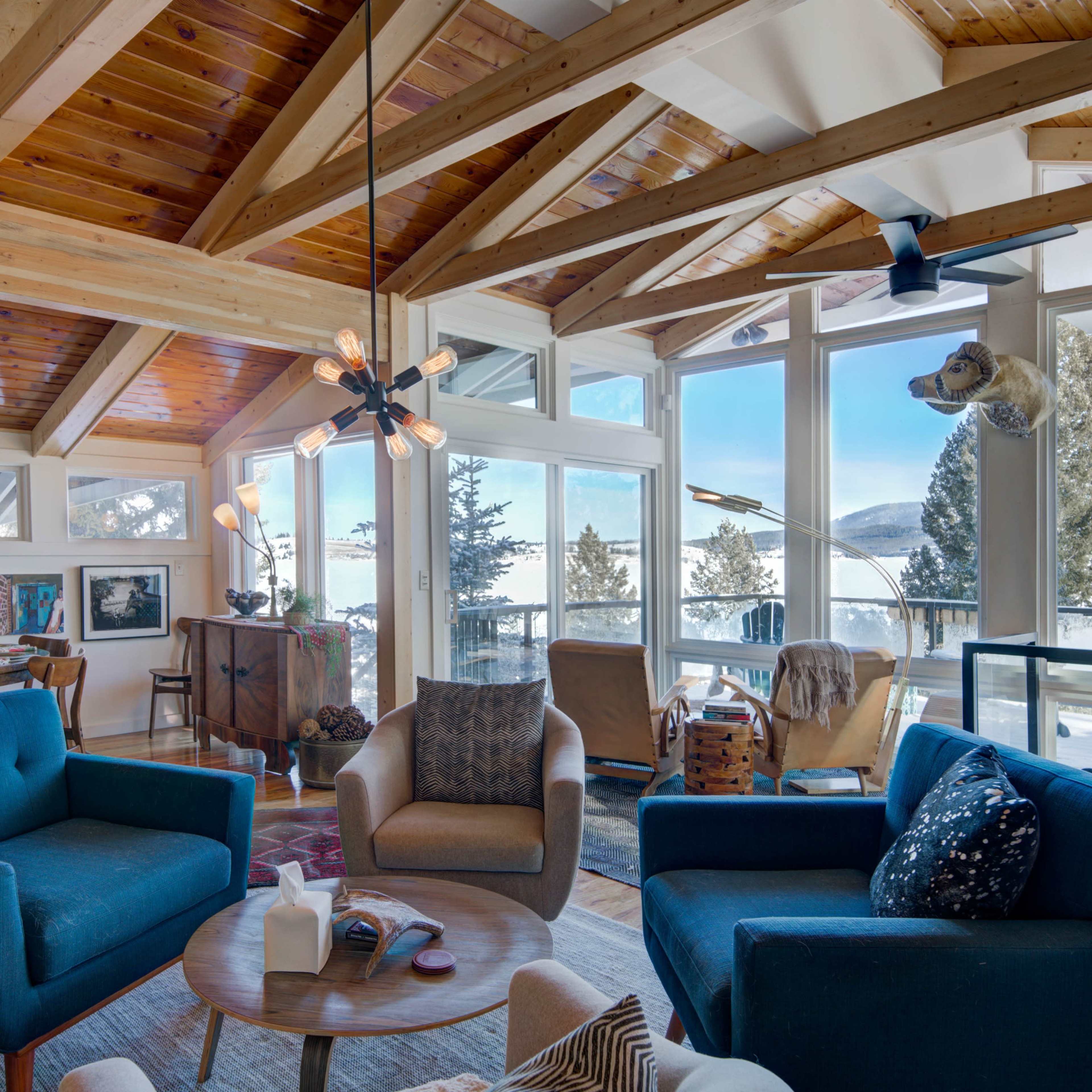 A spacious living room with modern furniture, large windows overlooking a snowy landscape, and exposed wooden beams on the ceiling.