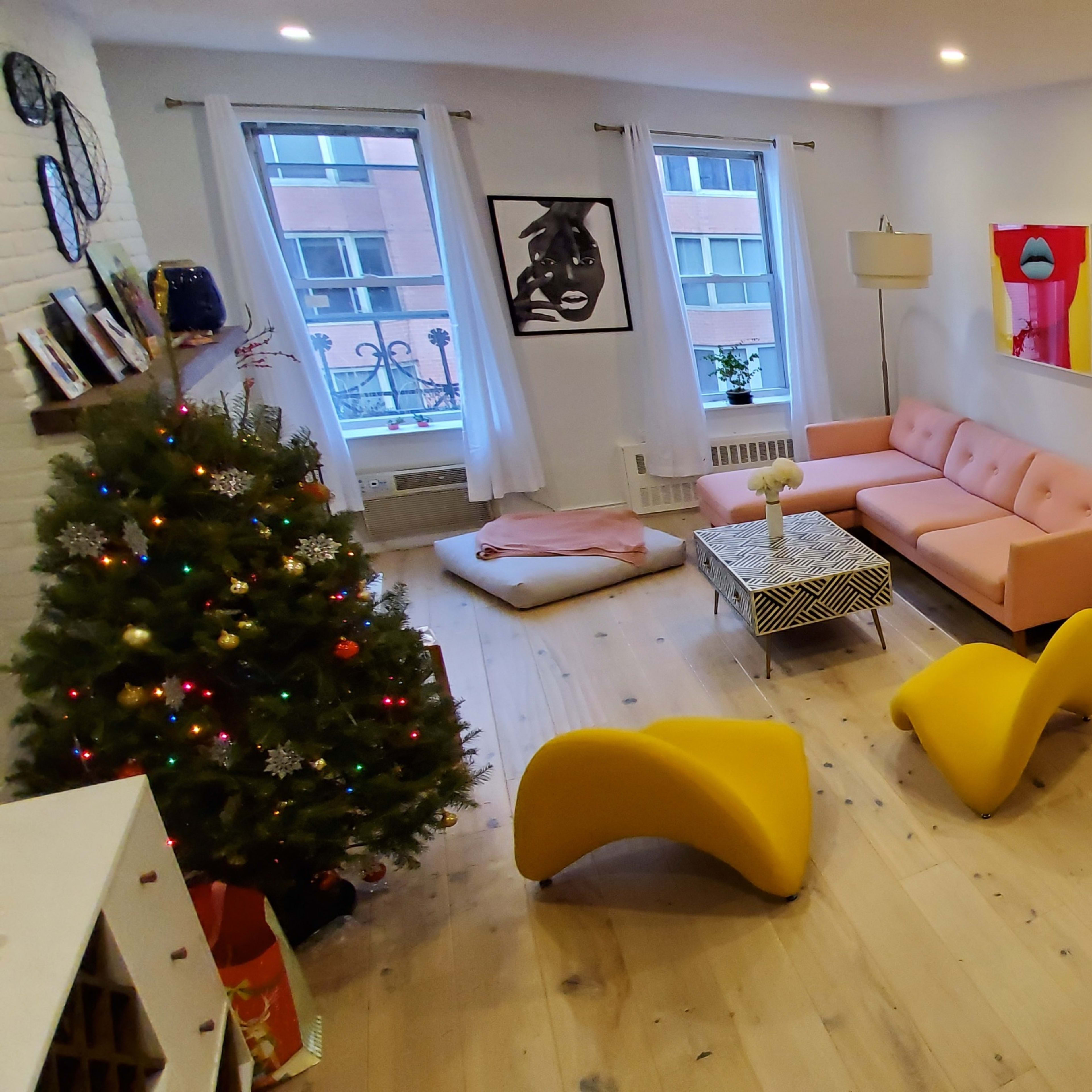 A modern living room with a pink sofa, yellow chairs, a small Christmas tree, and a dining area featuring black and white striped walls.