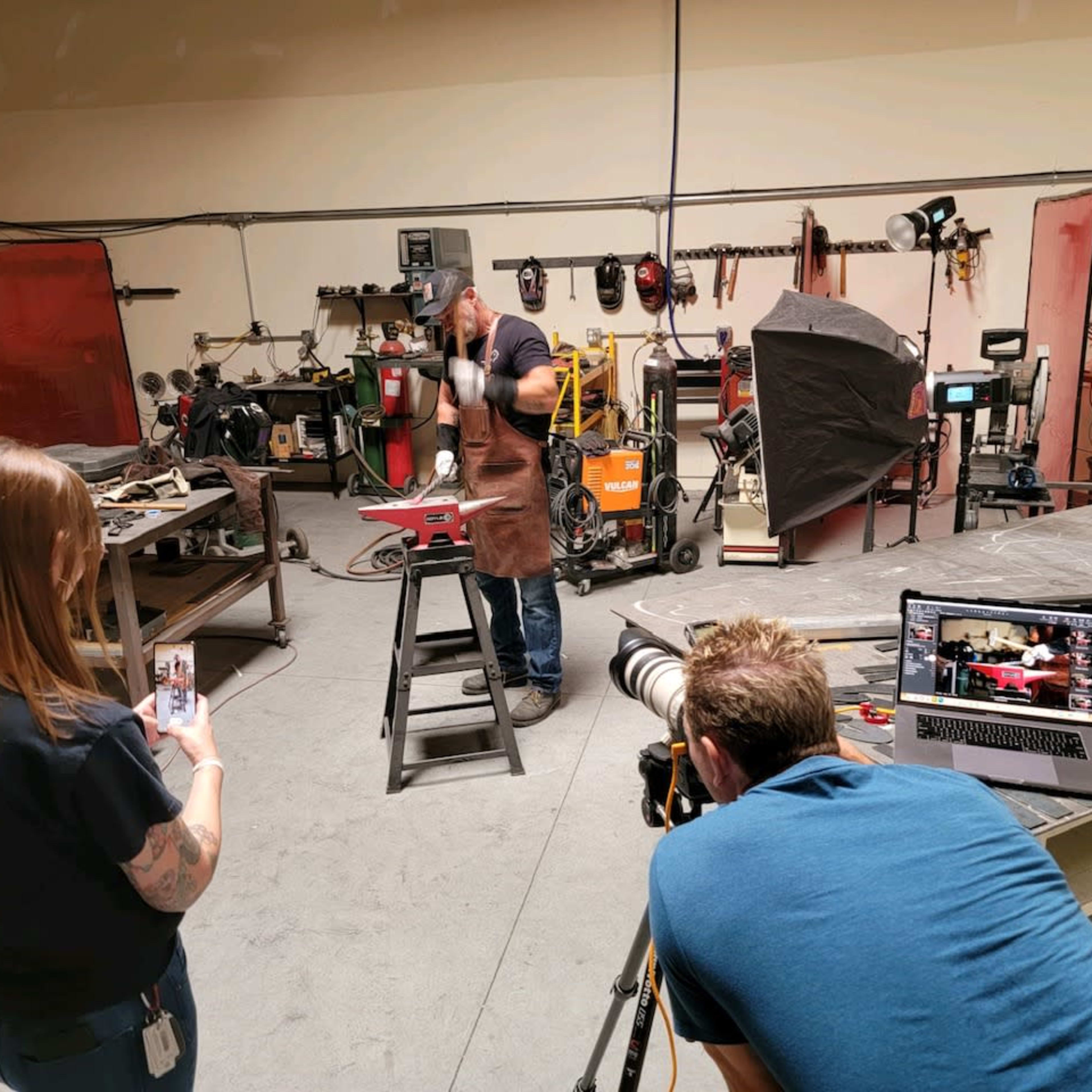 A person wearing an apron works on a metal project in a workshop while another person records on a phone and a third person operates a camera.