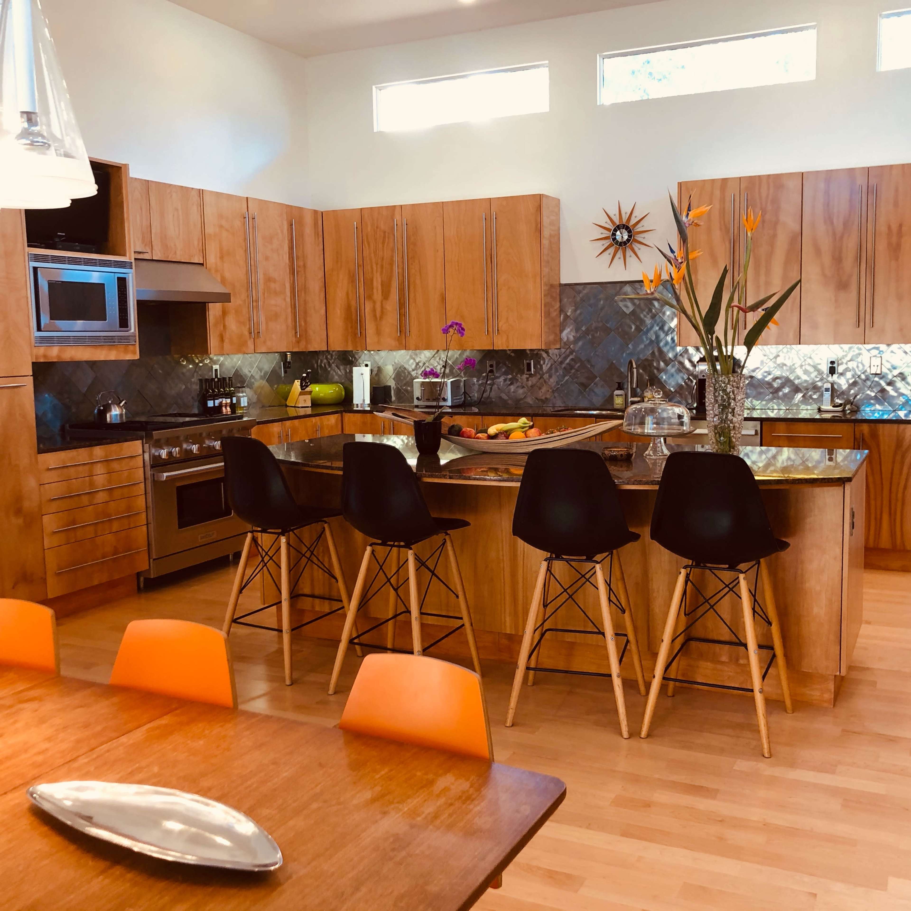A modern kitchen features wooden cabinetry, a central island with bar stools, and large windows allowing natural light.
