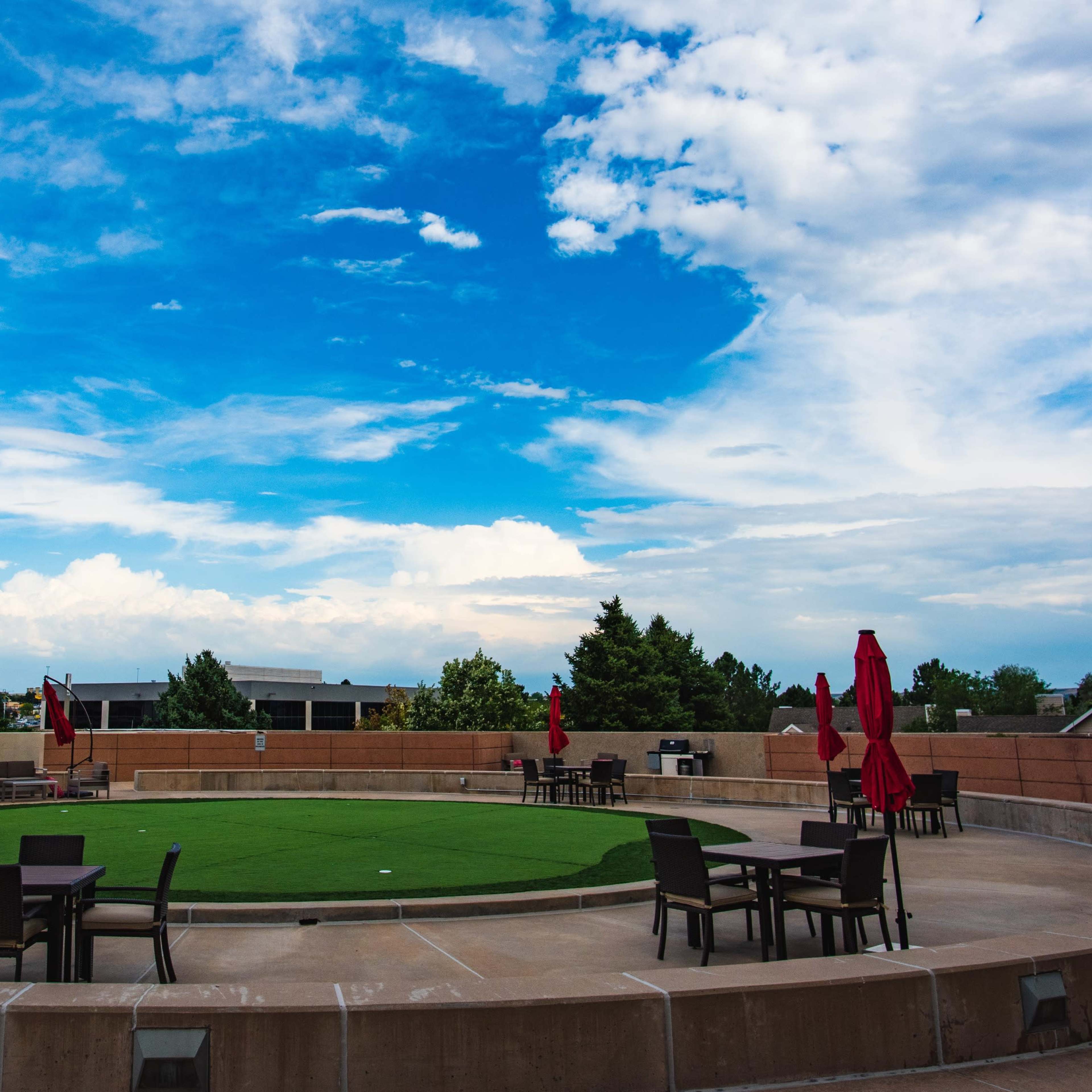 A circular plaza with green turf and scattered chairs under red umbrellas is set against a backdrop of a blue sky with scattered clouds.