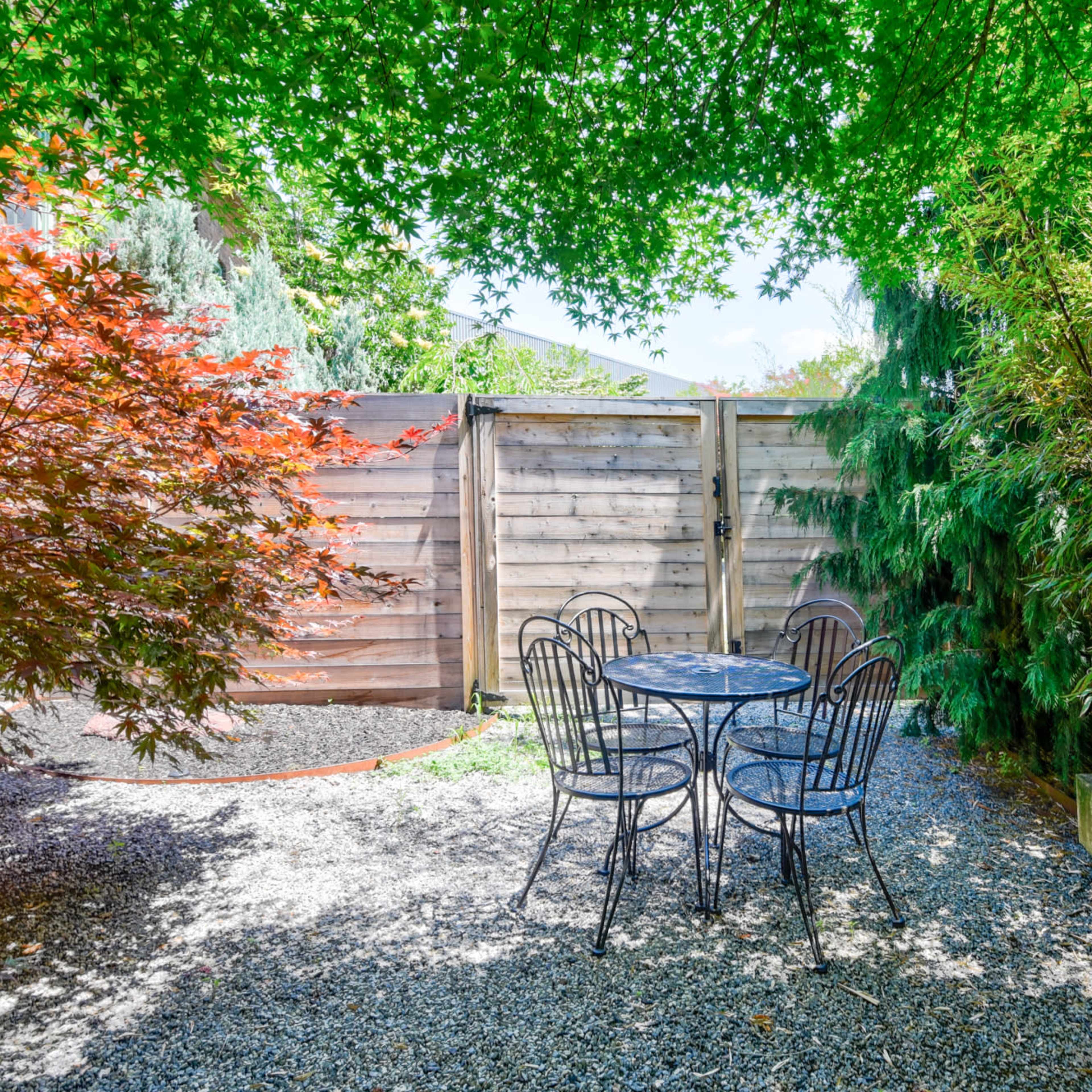 A small gravel patio features a round table and chairs surrounded by lush greenery and wooden fencing.