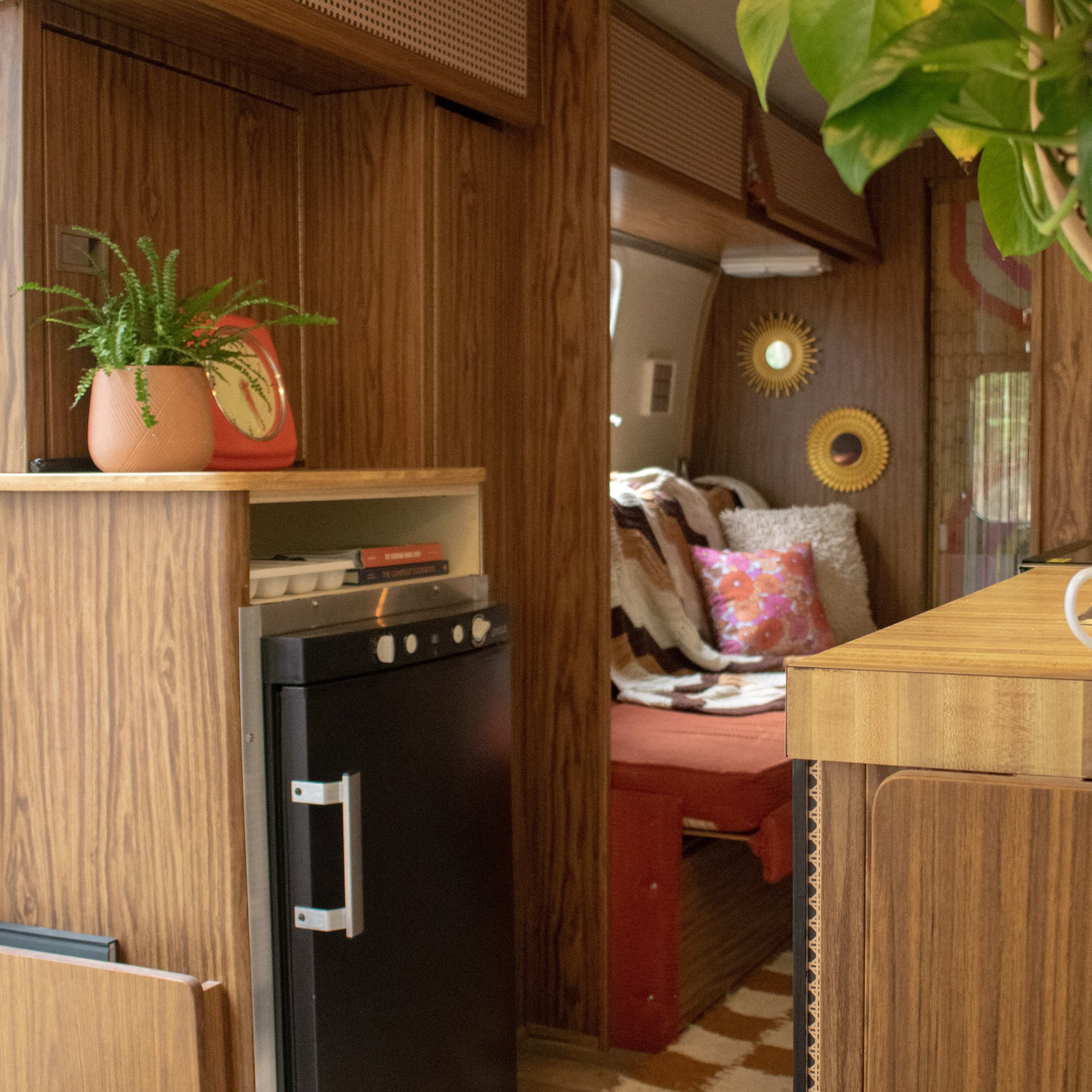 The interior of a cozy retro camper features wood paneling, a small kitchenette with a black fridge, and a seating area adorned with colorful cushions.