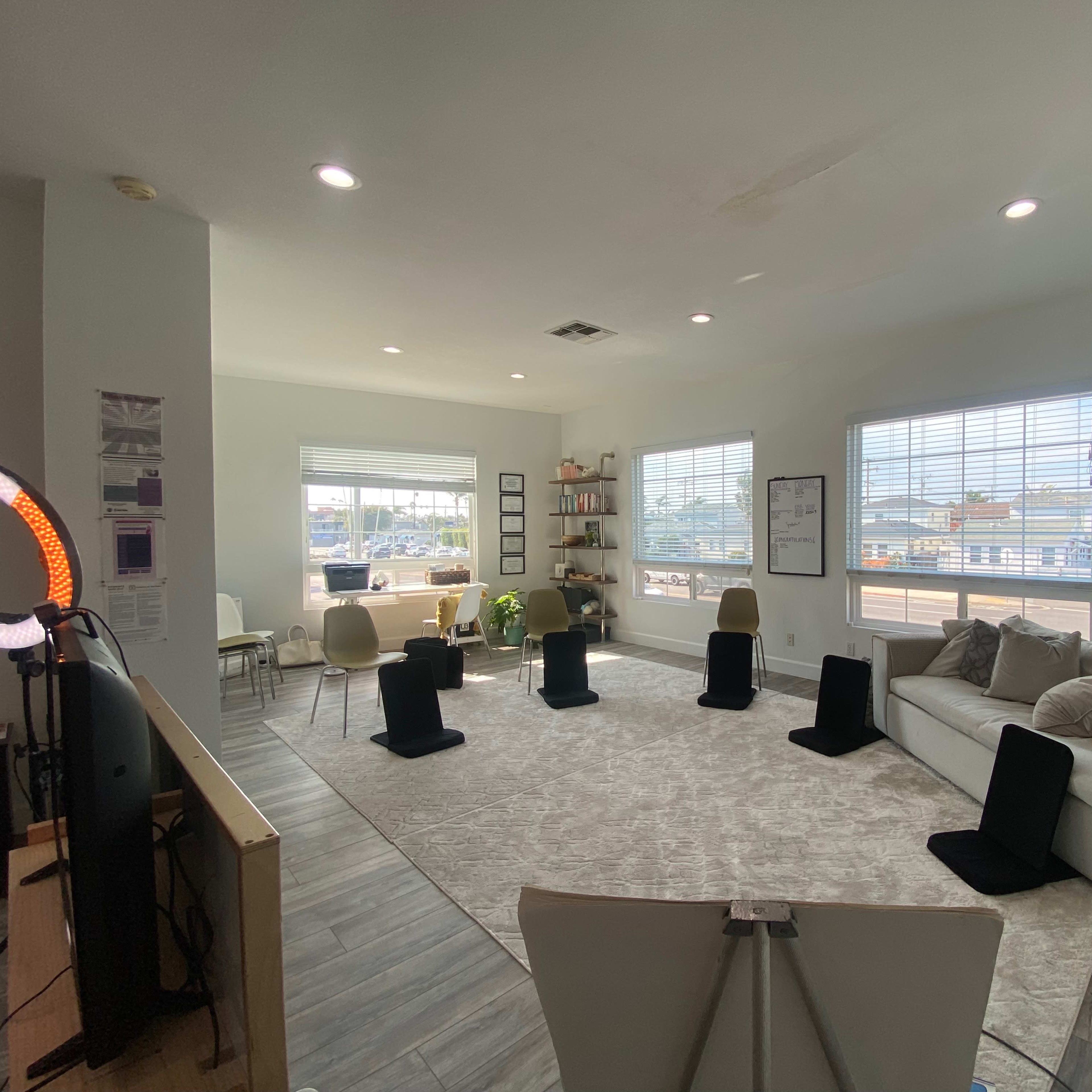 A spacious, well-lit living area featuring a large rug, a sectional couch, chairs, a bookshelf, and a desk with a view.