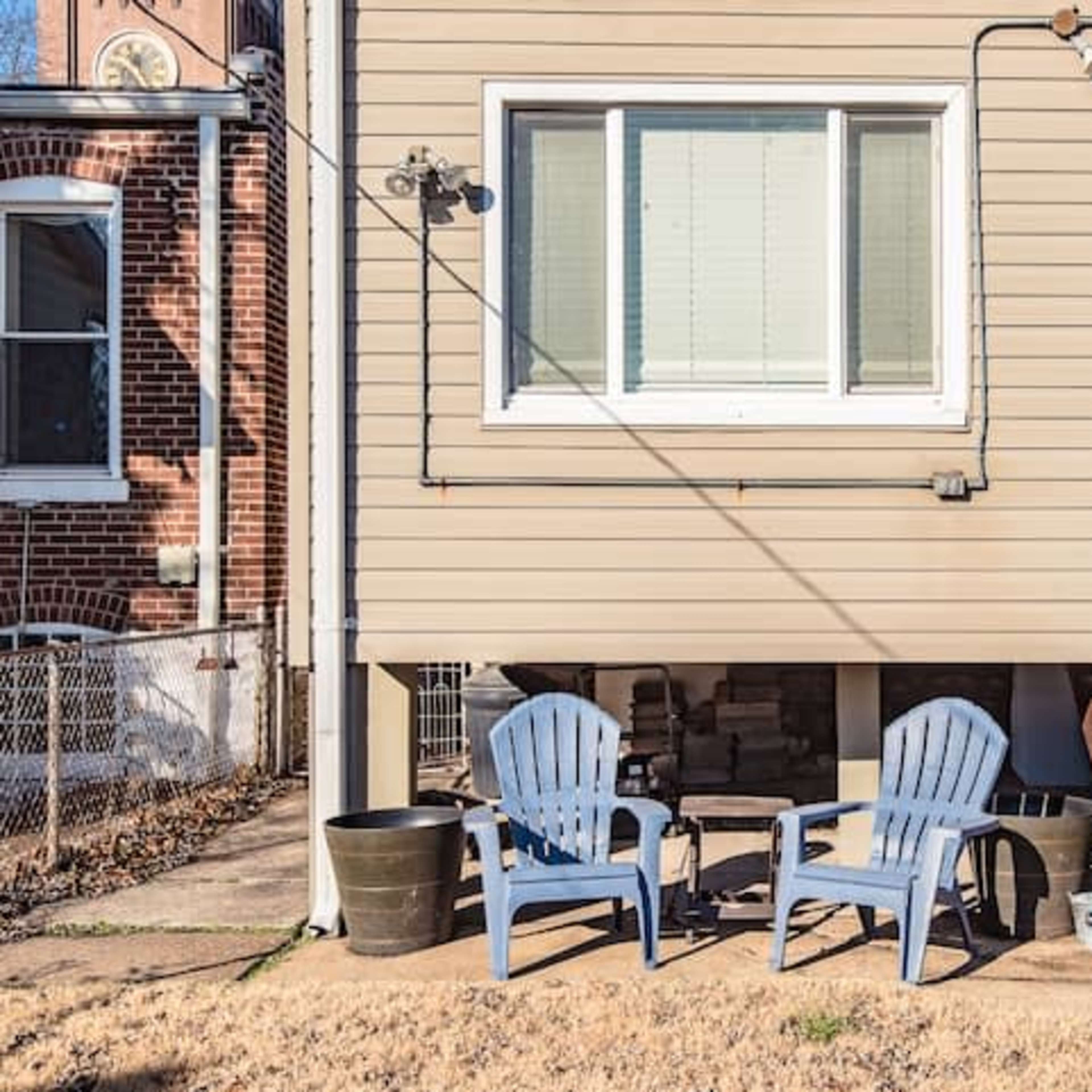 A small house with a staircase leading to a door, flanked by two blue Adirondack chairs on a grassy area.