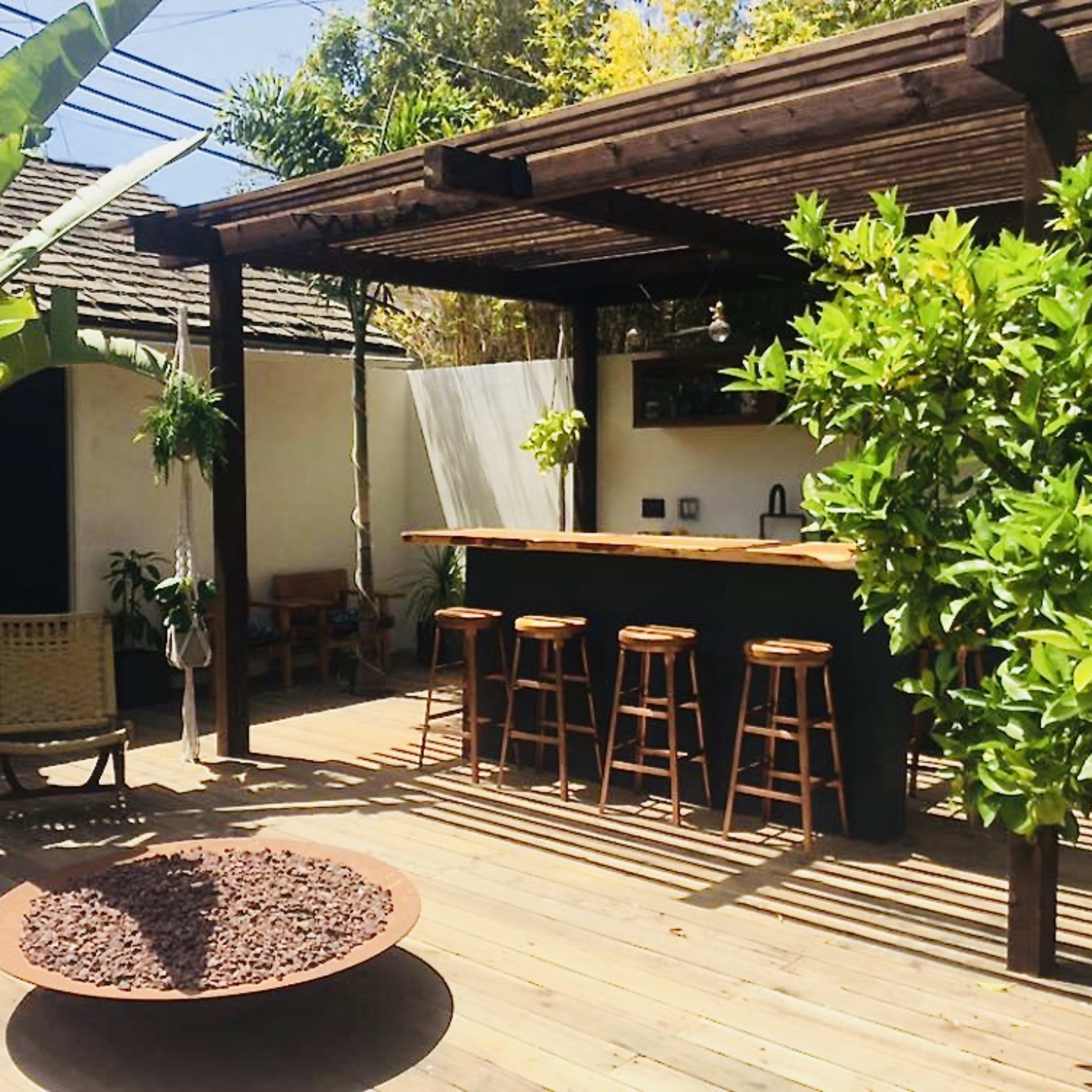 A wooden deck with outdoor seating, featuring a bar area and a circular fire pit surrounded by plants.
