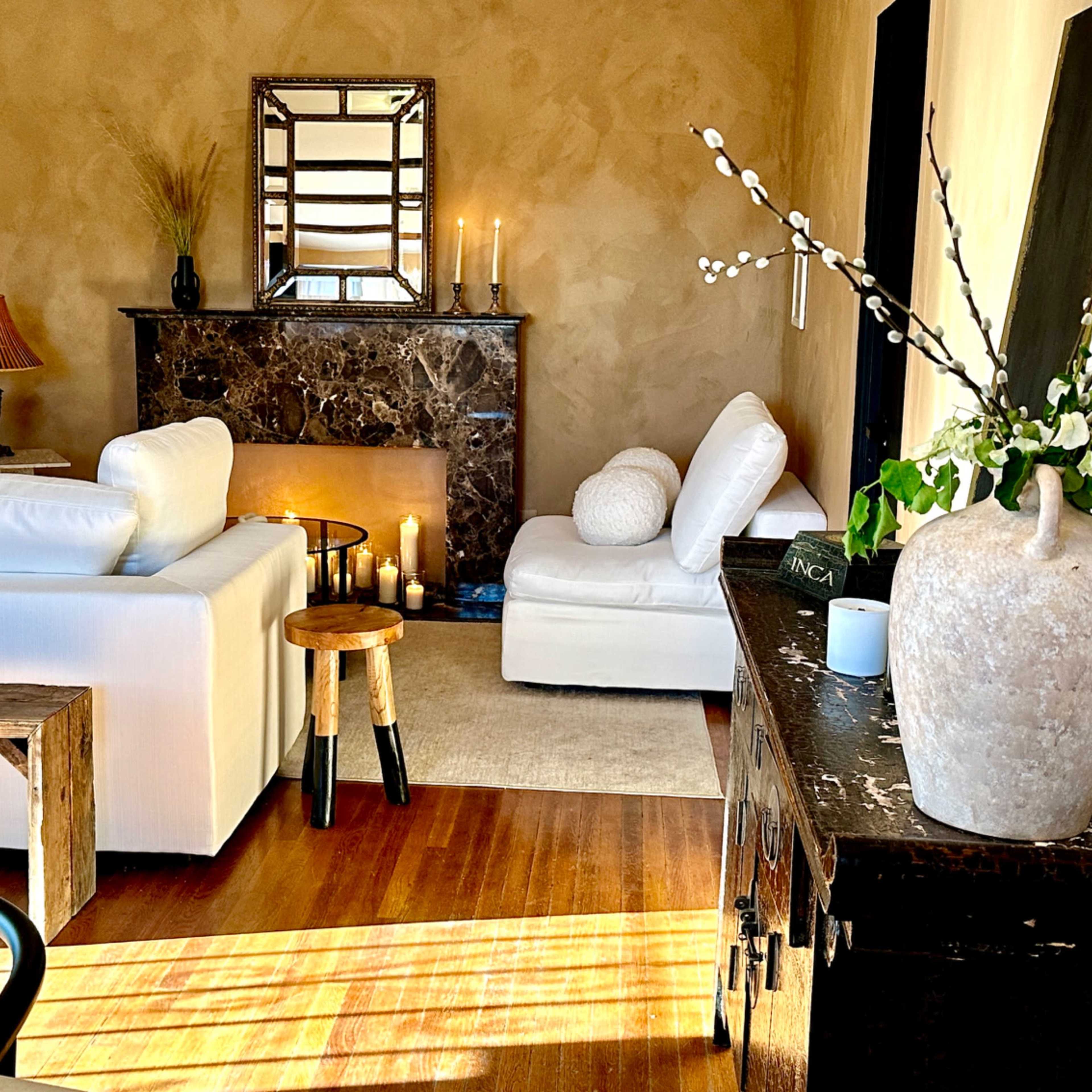 A neatly arranged living room with a white sofa, a marble fireplace adorned with candles, and a decoratively styled table featuring a vase and branches.