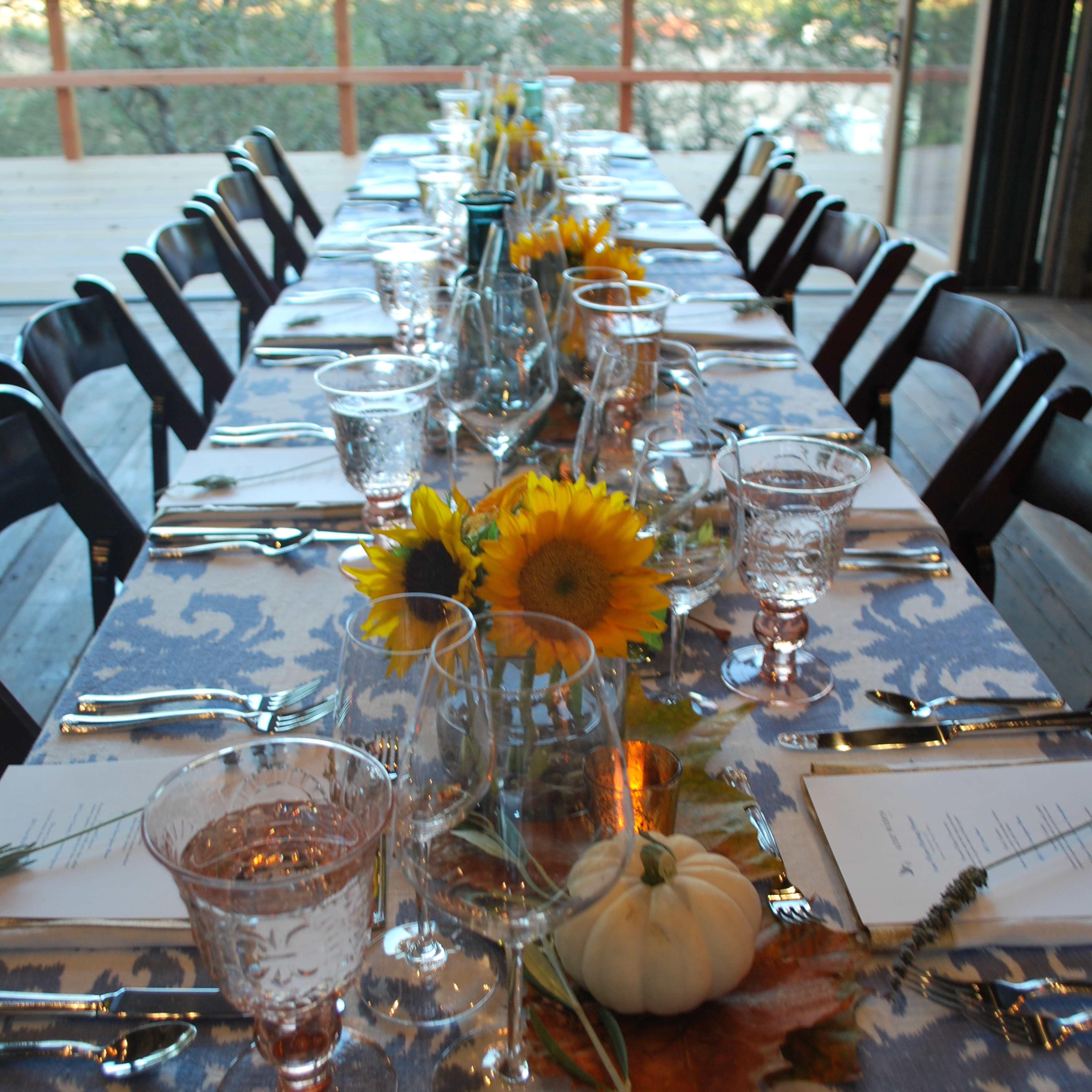 A long dining table is set with glasses, cutlery, and decorations including sunflowers and a small pumpkin.