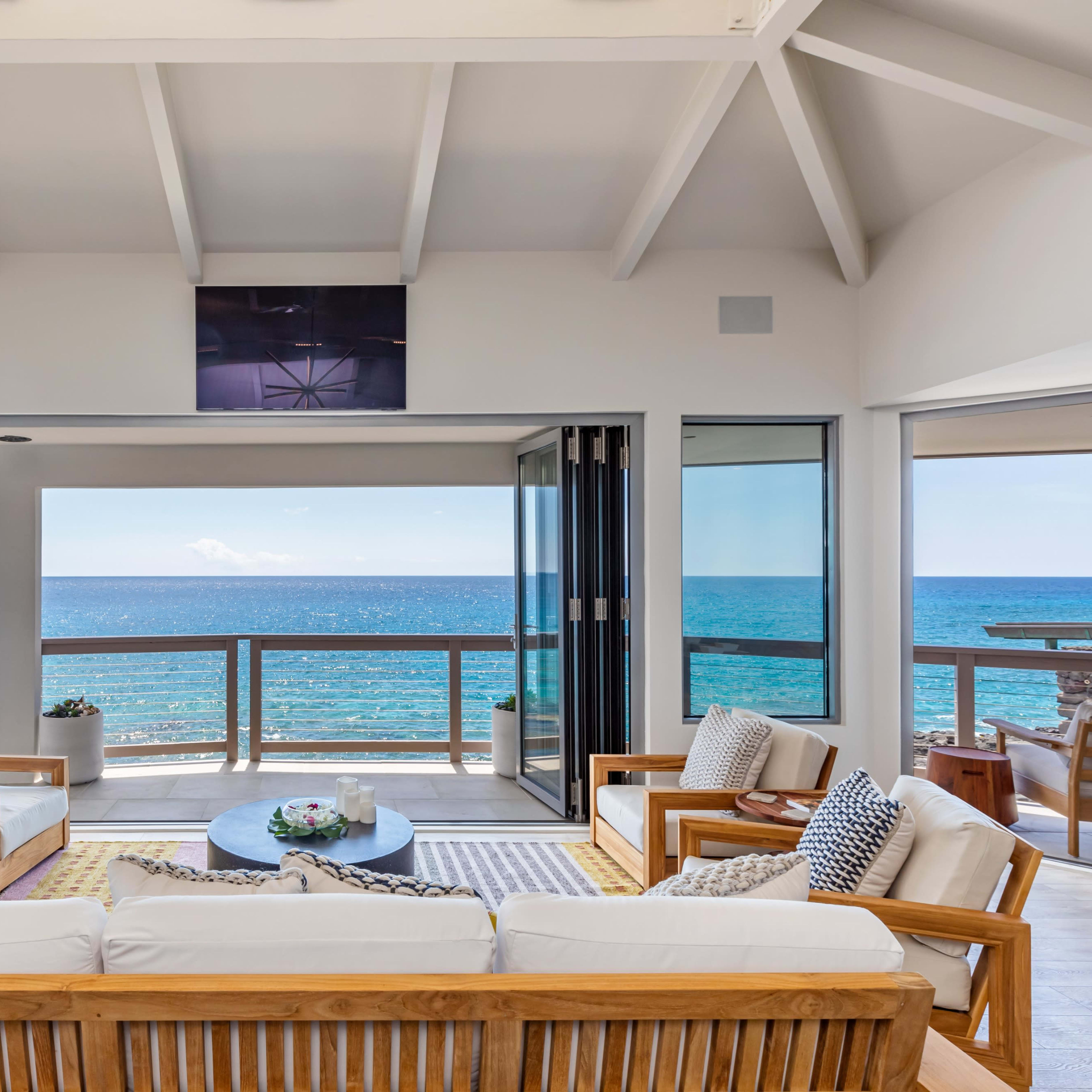 A coastal living room features large glass doors opening to a panoramic ocean view, with wooden furniture arranged around a central coffee table.