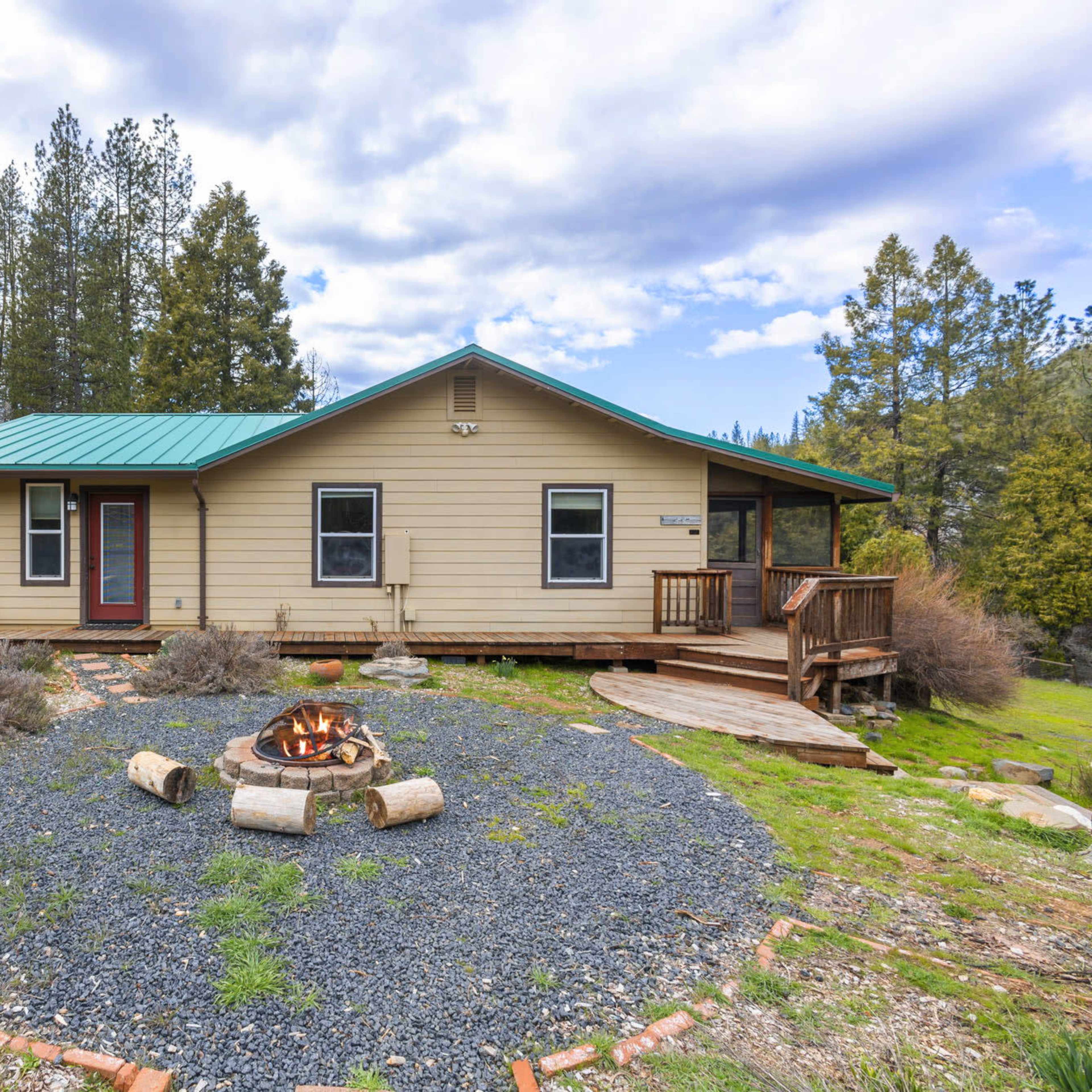 A single-story house with a green metal roof is situated near a circular fire pit surrounded by gravel and landscaping in a wooded area.