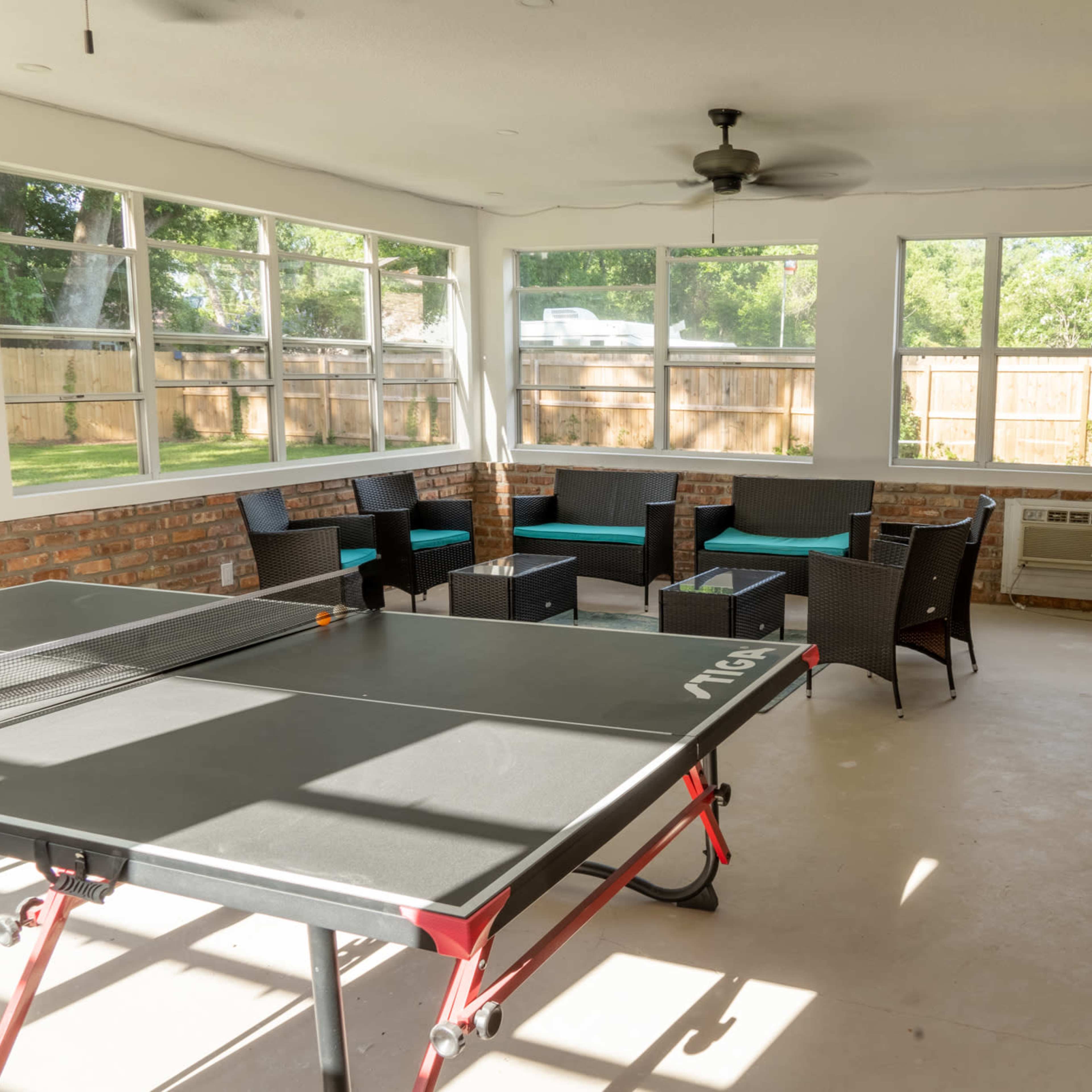 A ping pong table is placed in a sunlit room with large windows, surrounded by seating and brick walls.