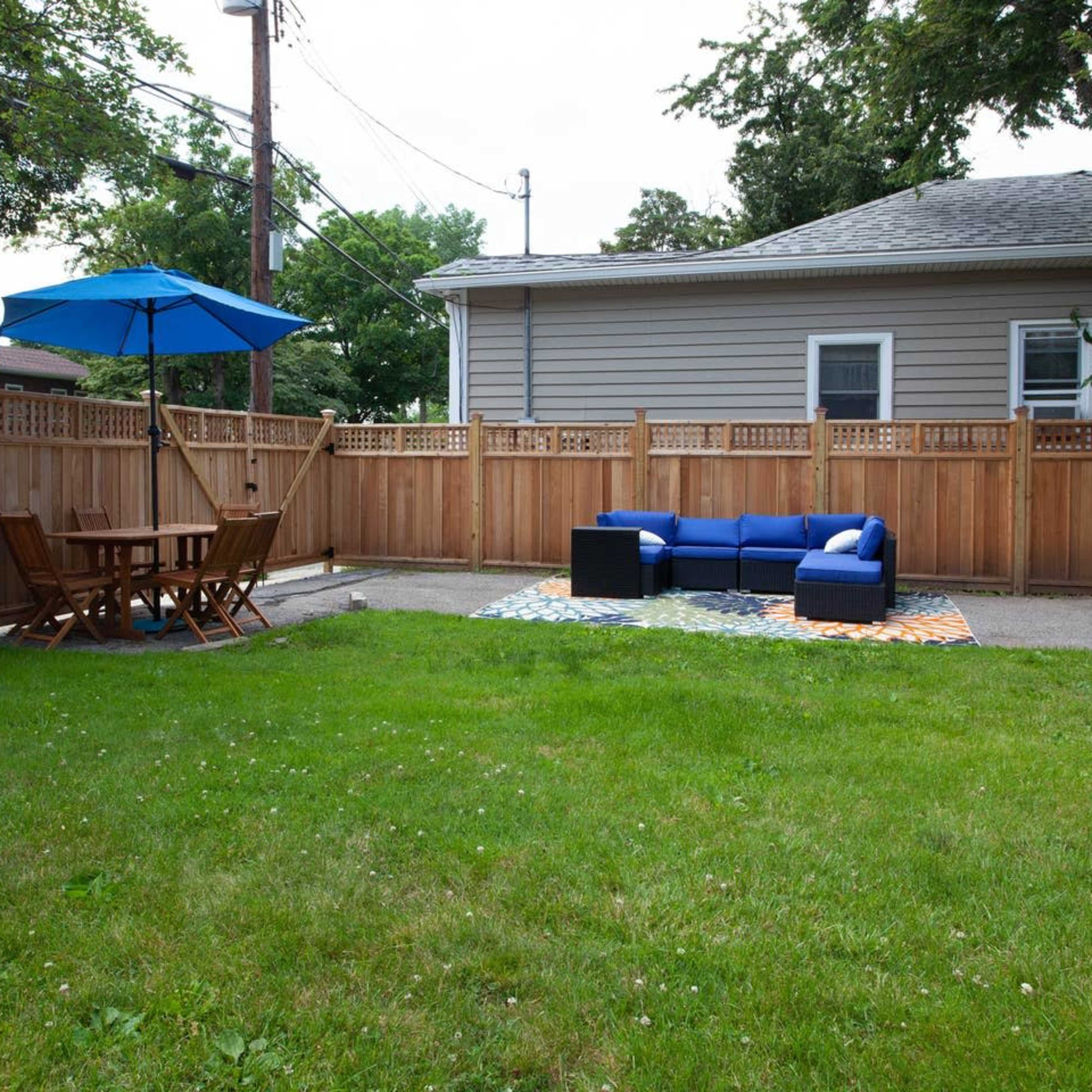 A backyard with a grassy area, a blue outdoor couch, a wooden dining table with chairs, and an umbrella, all enclosed by a wooden fence.