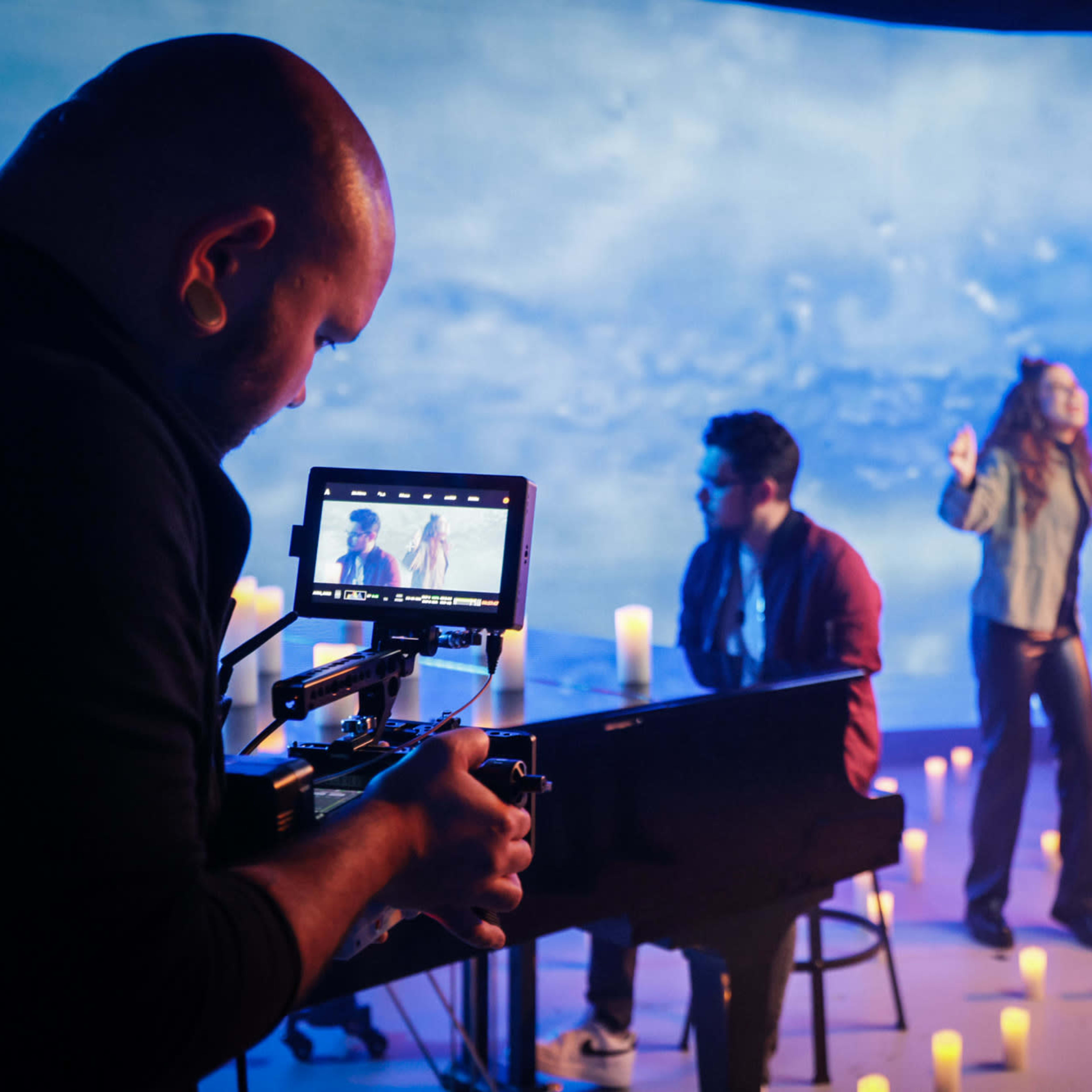 A musician performs in front of a blue backdrop while a camera operator captures the scene, surrounded by lit candles.