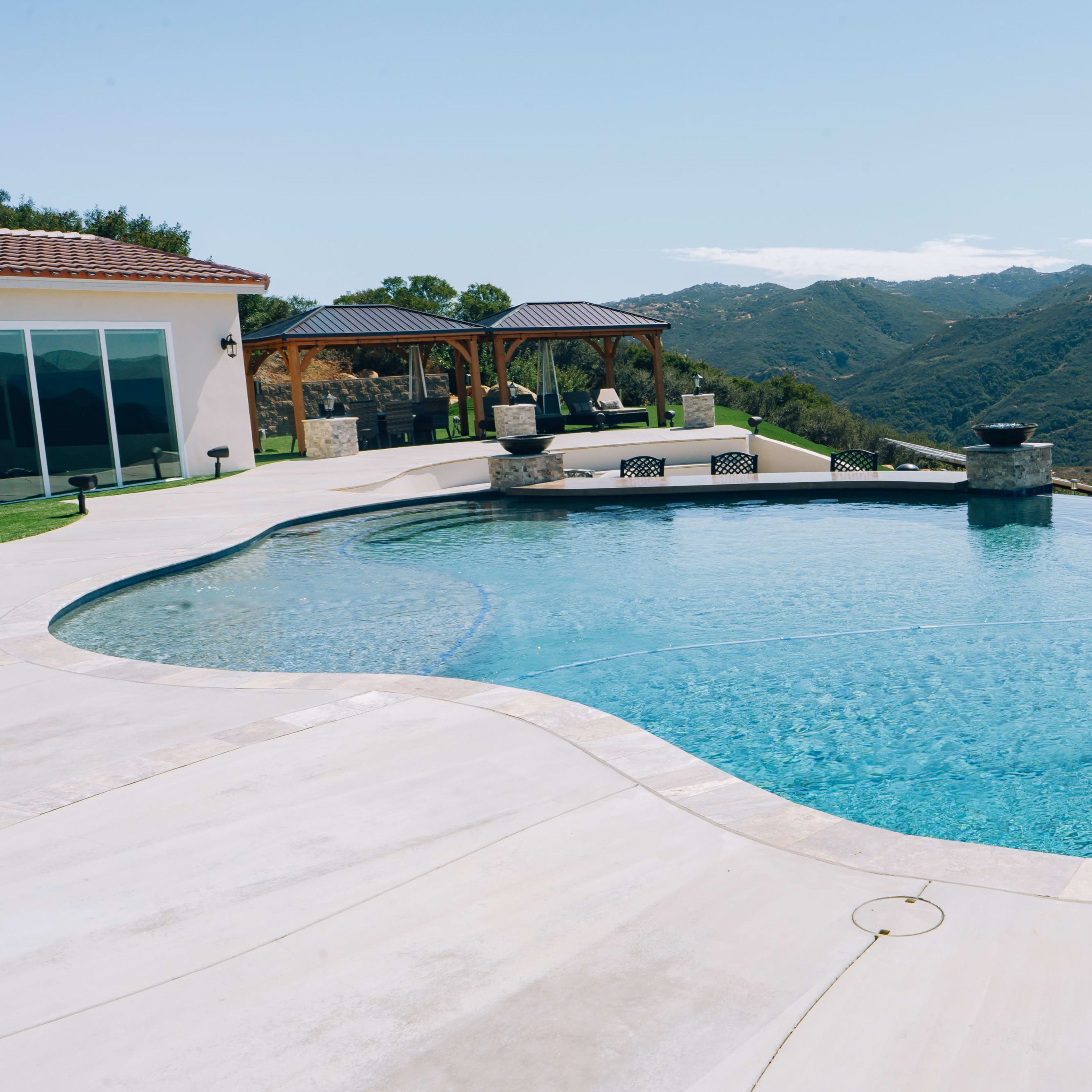 A modern pool overlooking a mountainous landscape, with a patio and seating areas nearby.