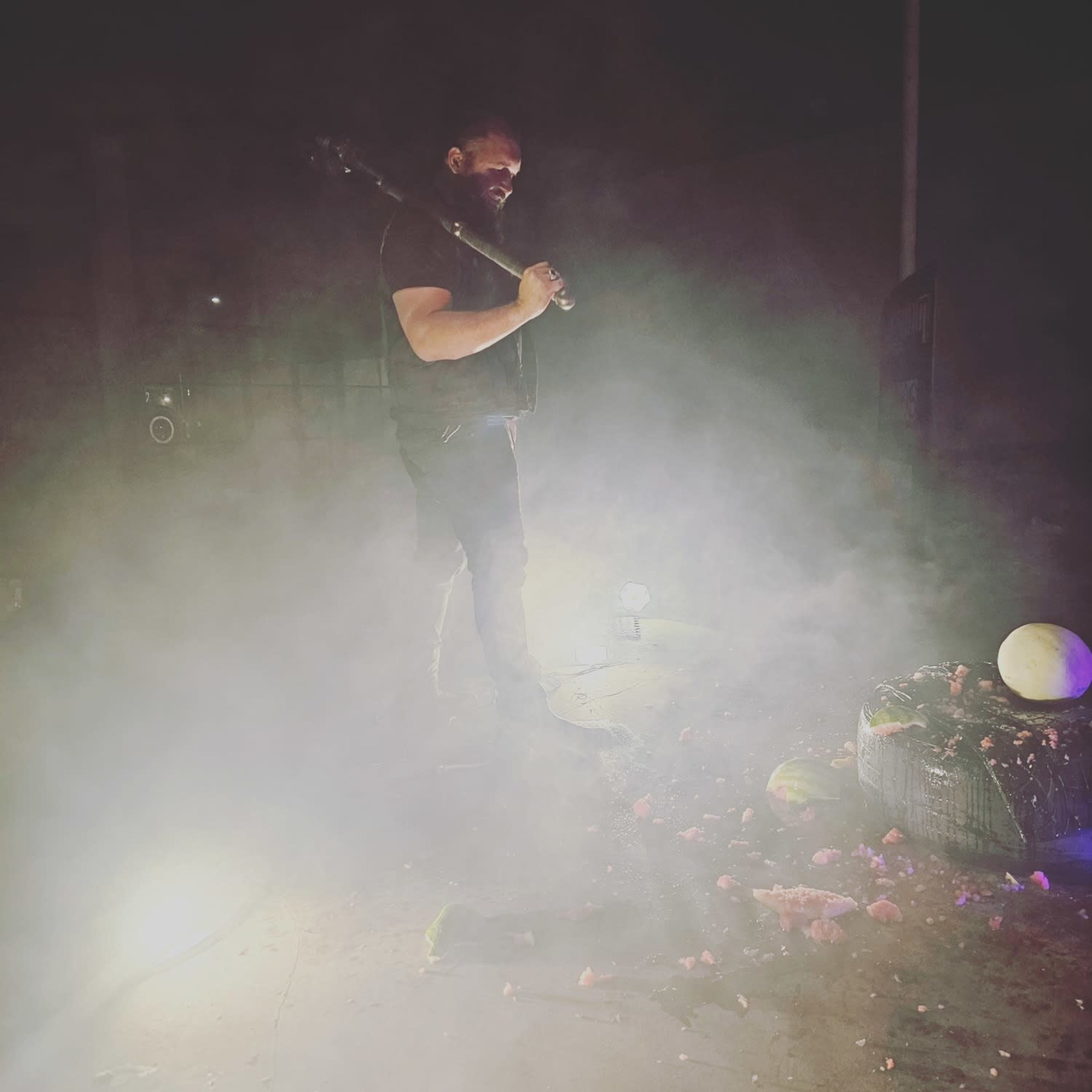 A person holding a baseball bat stands over a smashed watermelon in a smoky environment illuminated by lights.