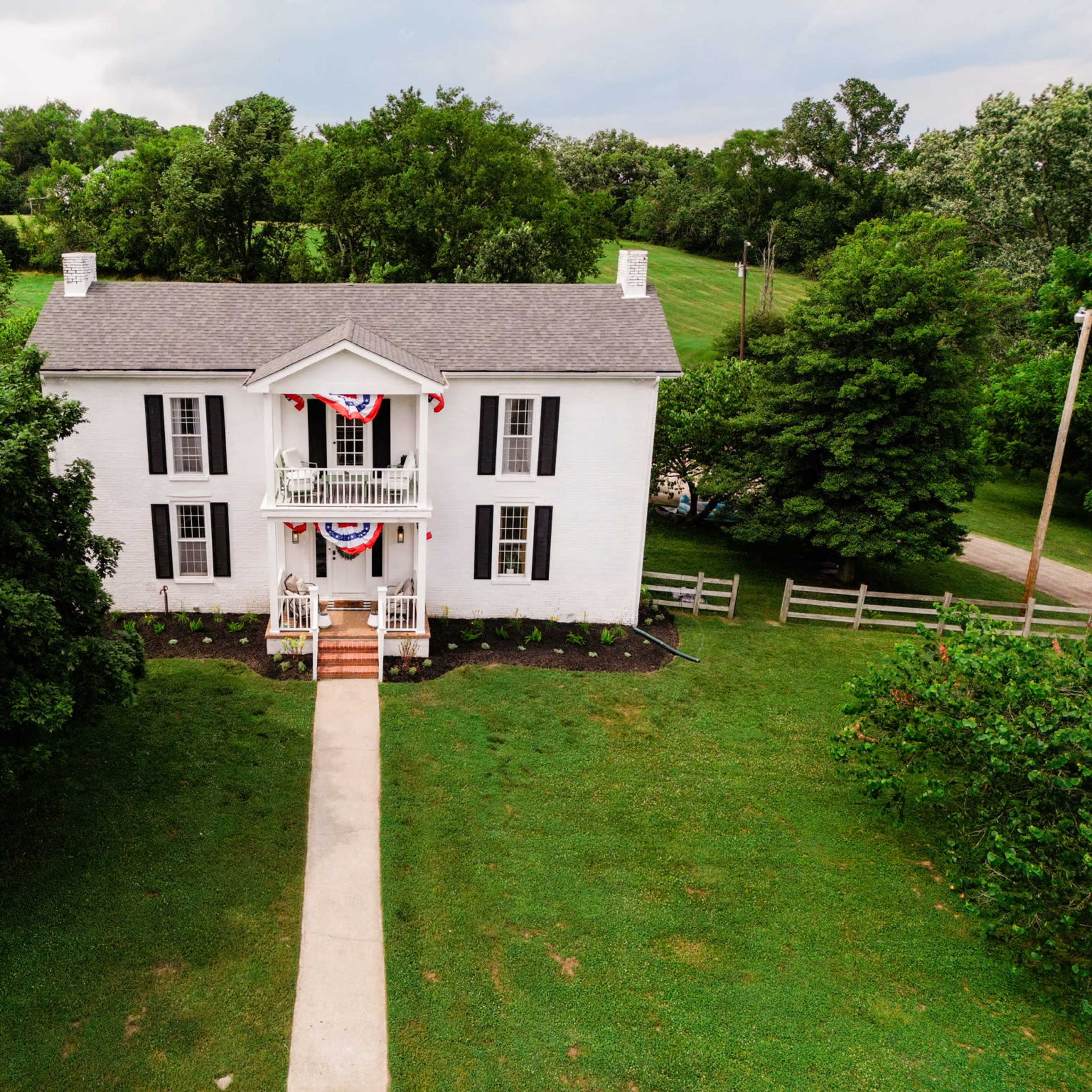 A white two-story house surrounded by trees, with red, white, and blue bunting displayed on the front porch.