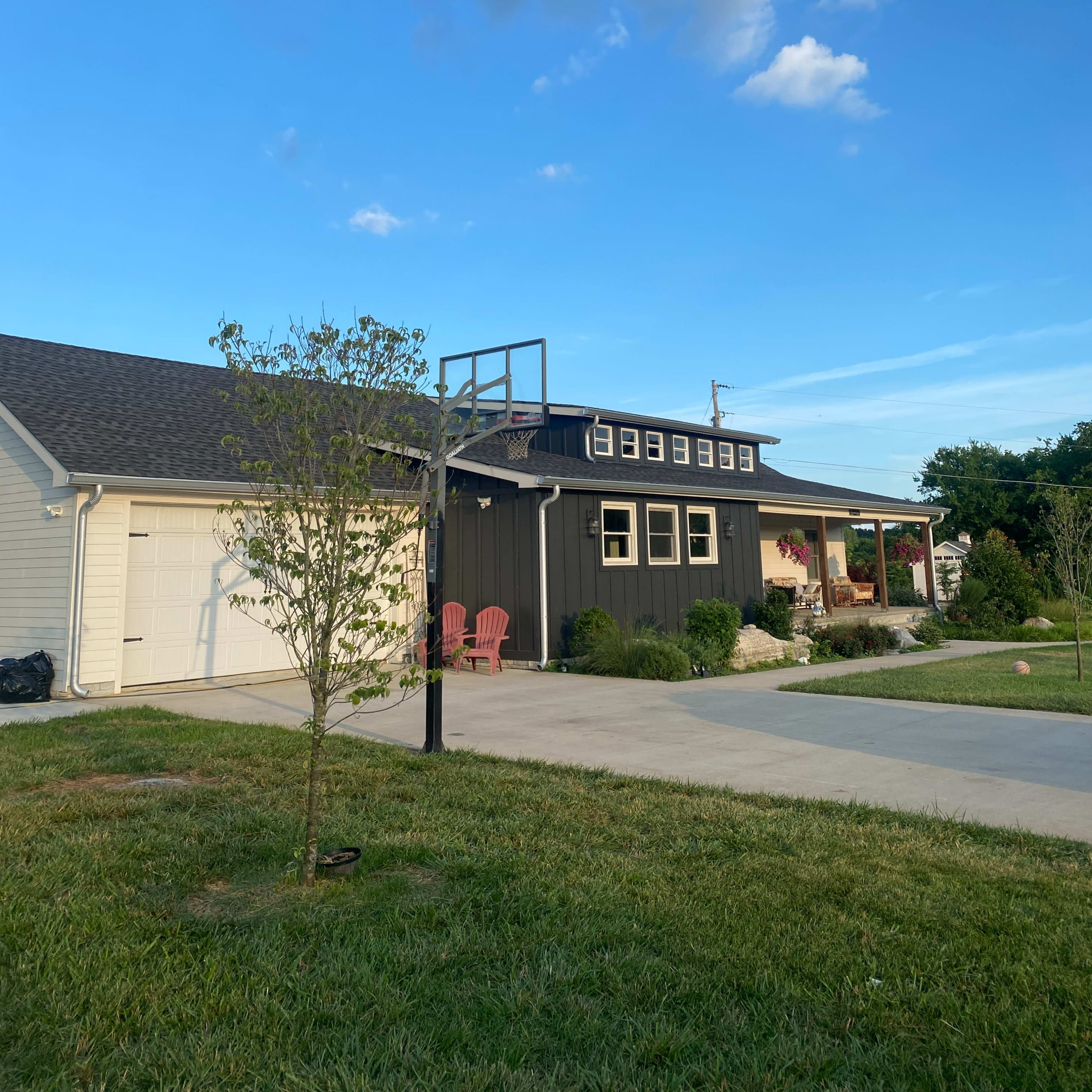 A two-story black and white house with a basketball hoop stands next to a driveway lined with green grass under a blue sky with scattered clouds.