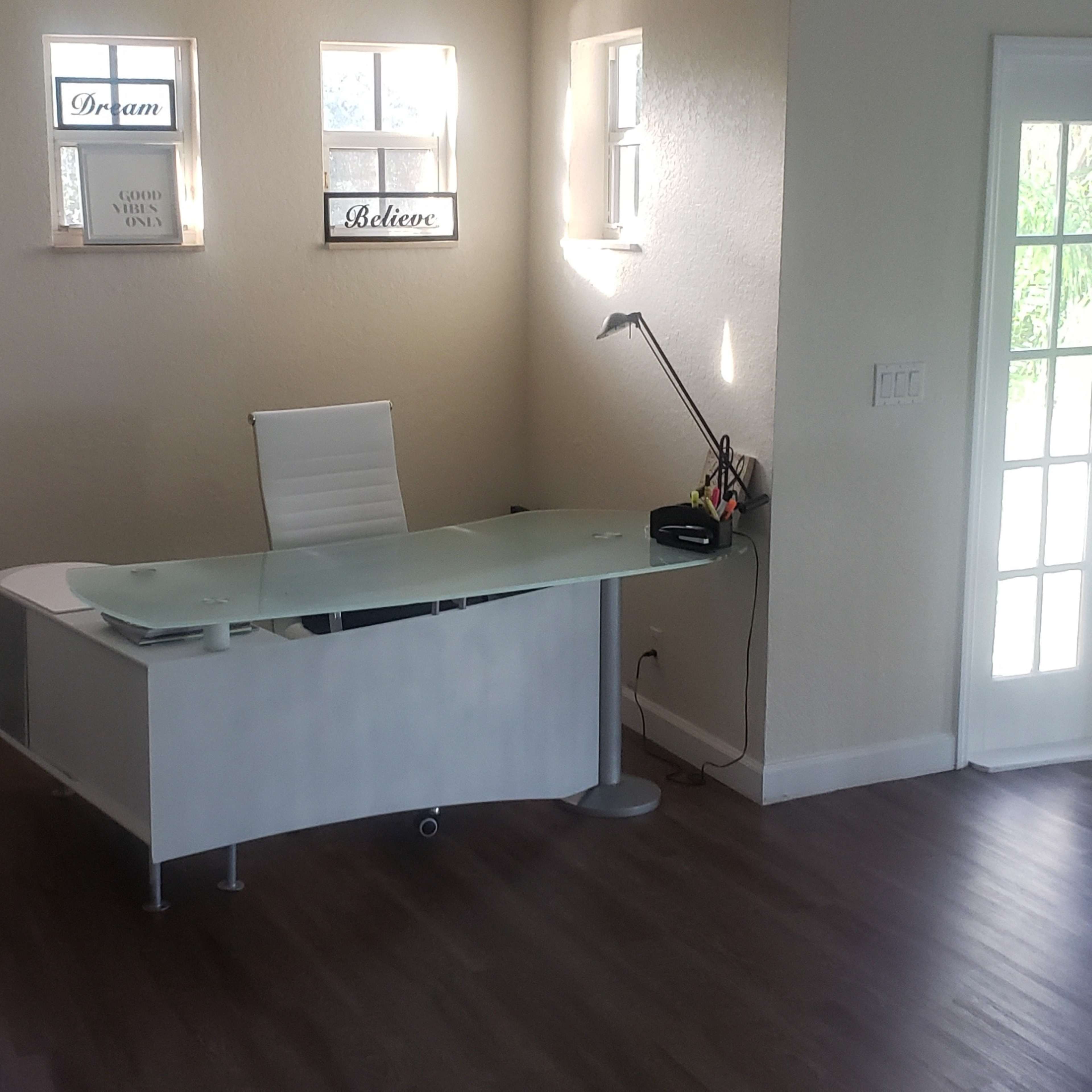 A modern office space with a glass desk, a chair, a bookshelf, and large windows allowing light to enter.
