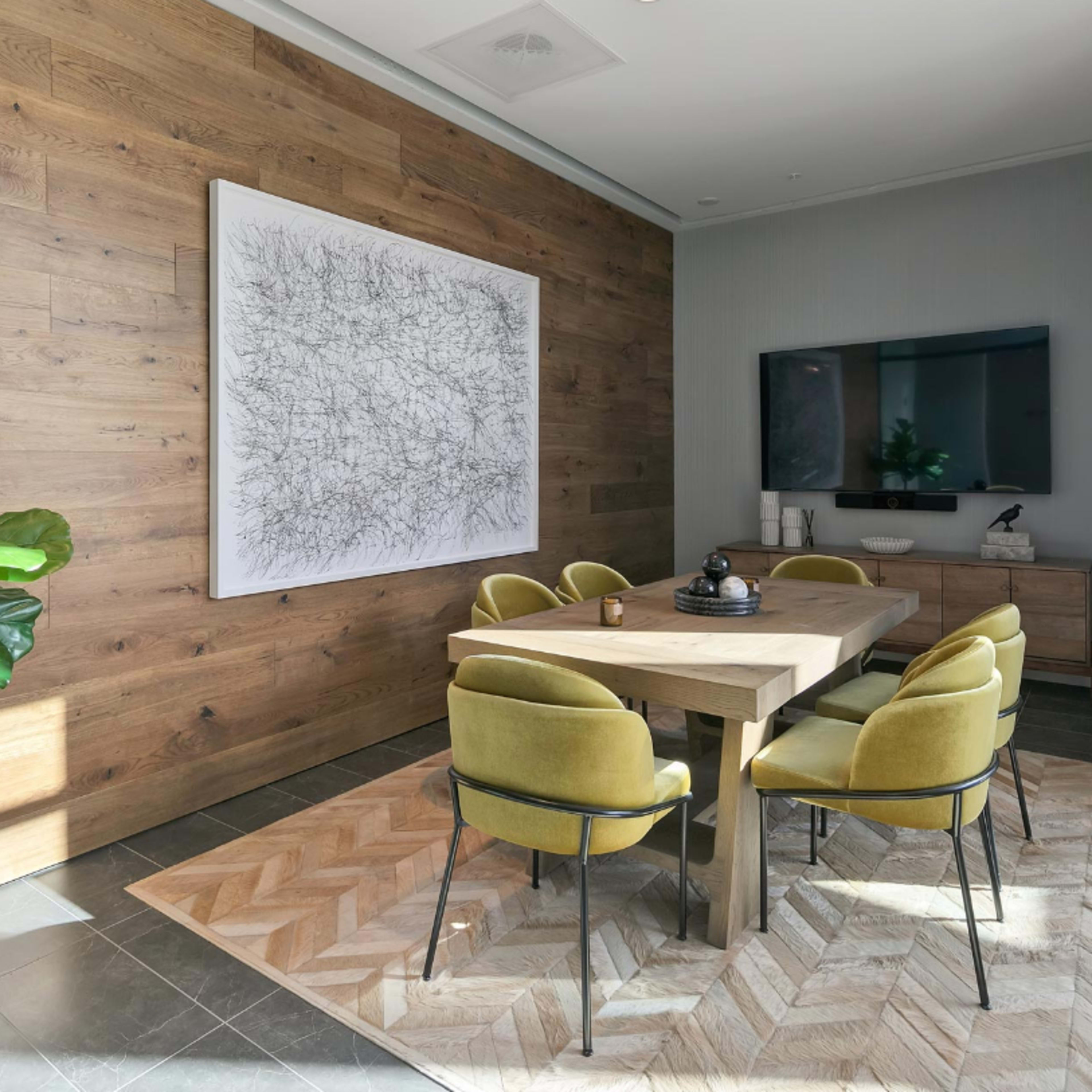 A modern dining room features a wooden table surrounded by yellow chairs, a large abstract artwork on the wall, and a television mounted above a side cabinet.