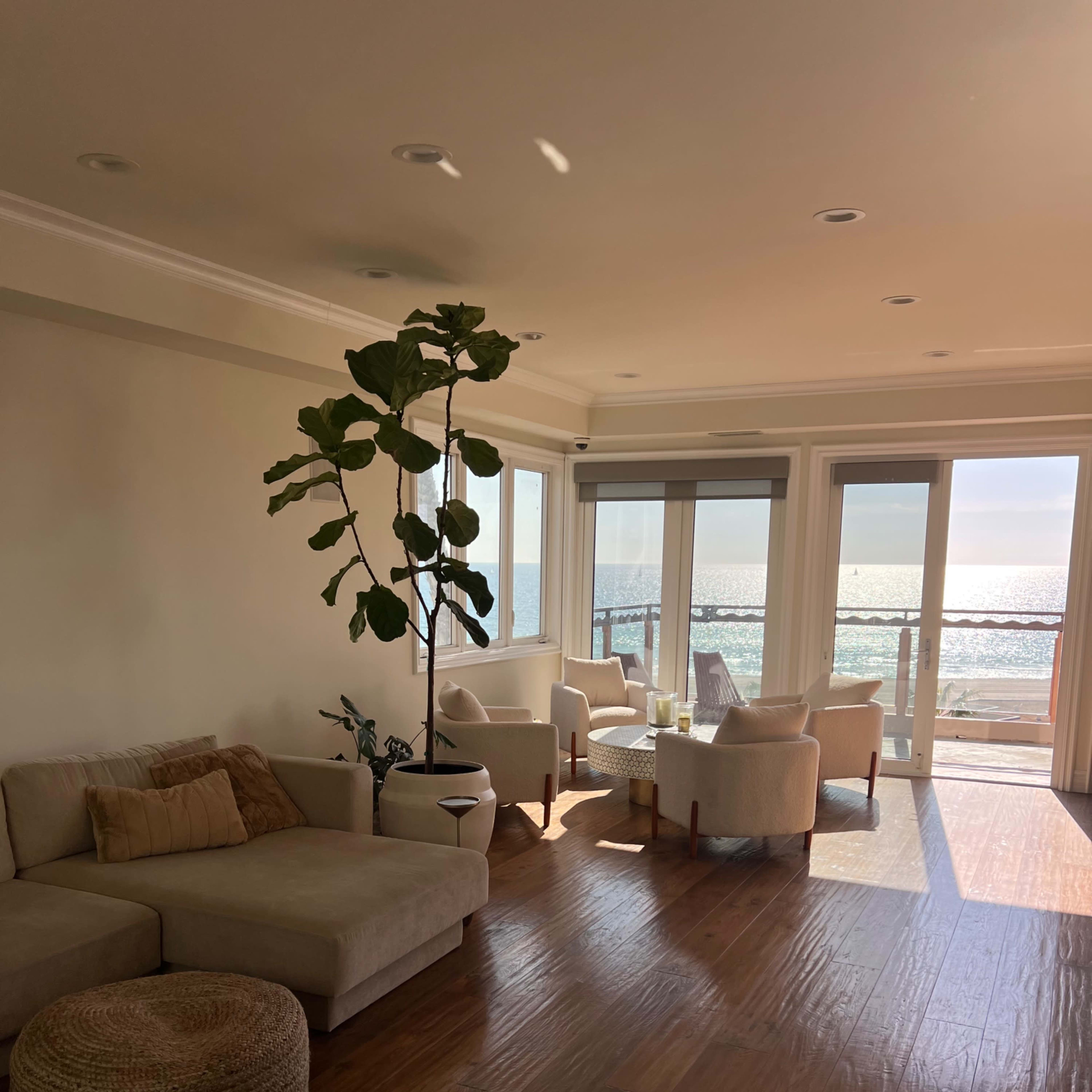 A spacious living room features a light-colored sofa, a round ottoman, and several chairs arranged near large windows overlooking the ocean.