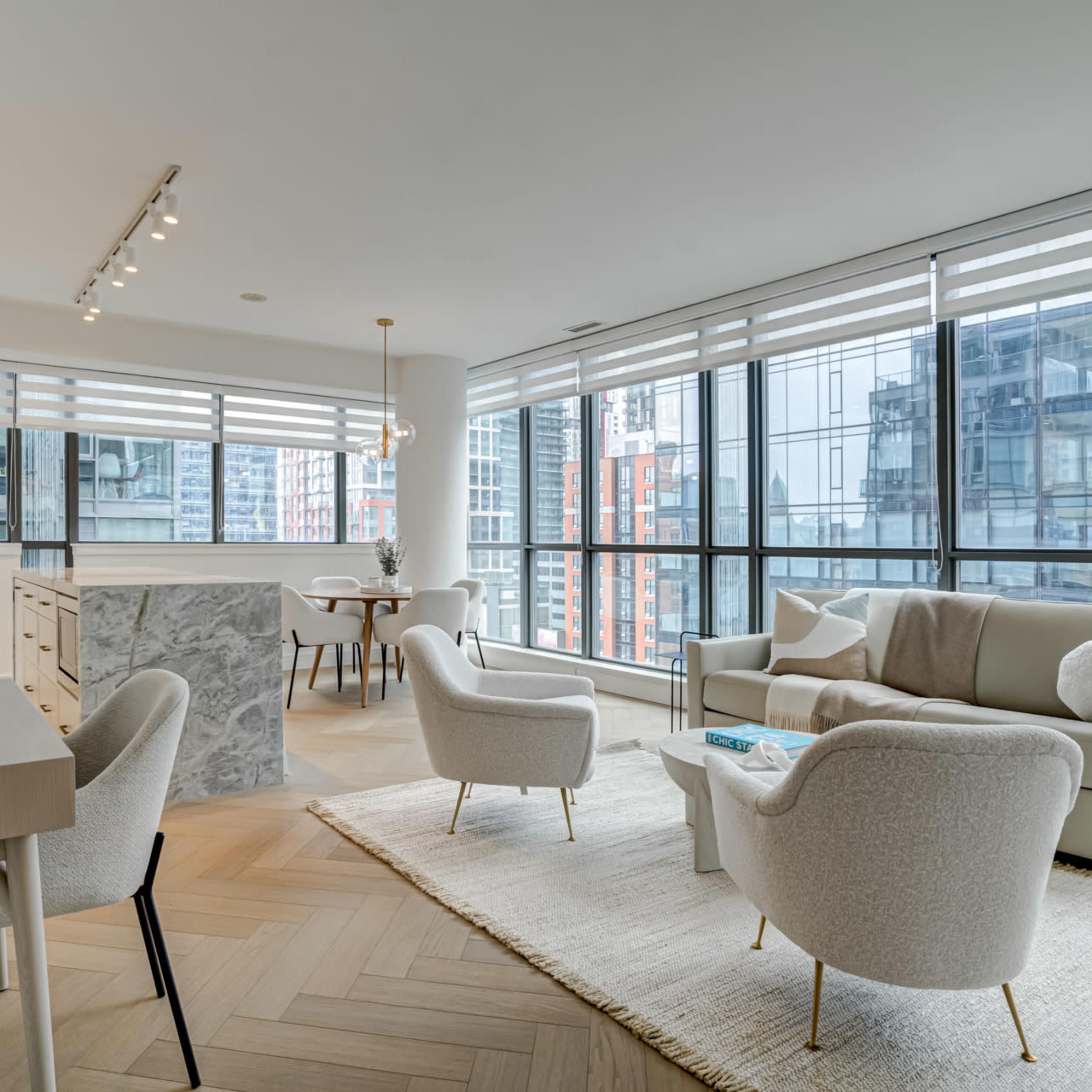 A modern living room features a light-colored sofa, two armchairs, and large windows overlooking city buildings.