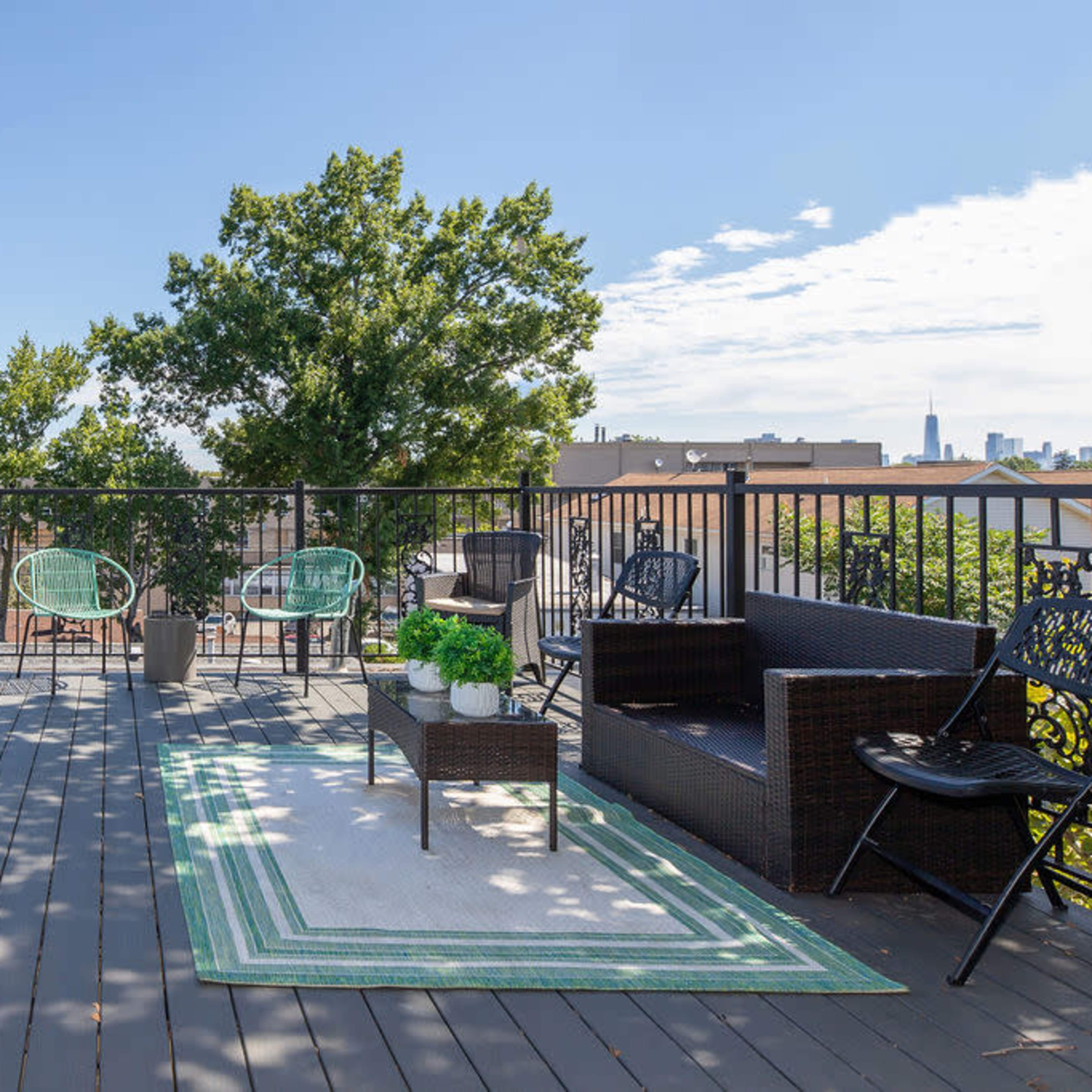 A rooftop terrace with various seating options and a striped rug, surrounded by trees and buildings in the background.