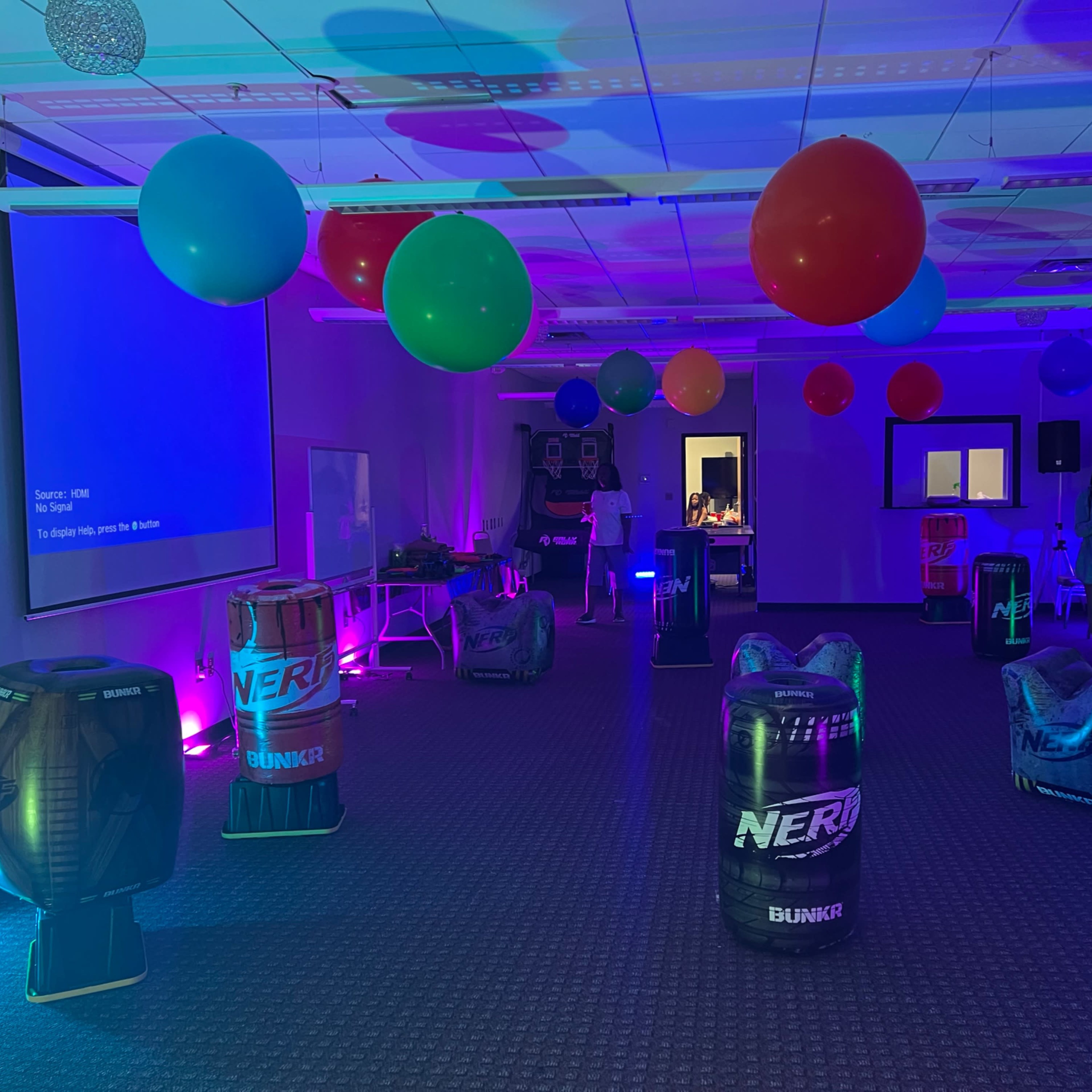The room is set up for an event, featuring colorful balloons and a series of Nerf-themed obstacles arranged around a carpeted floor.