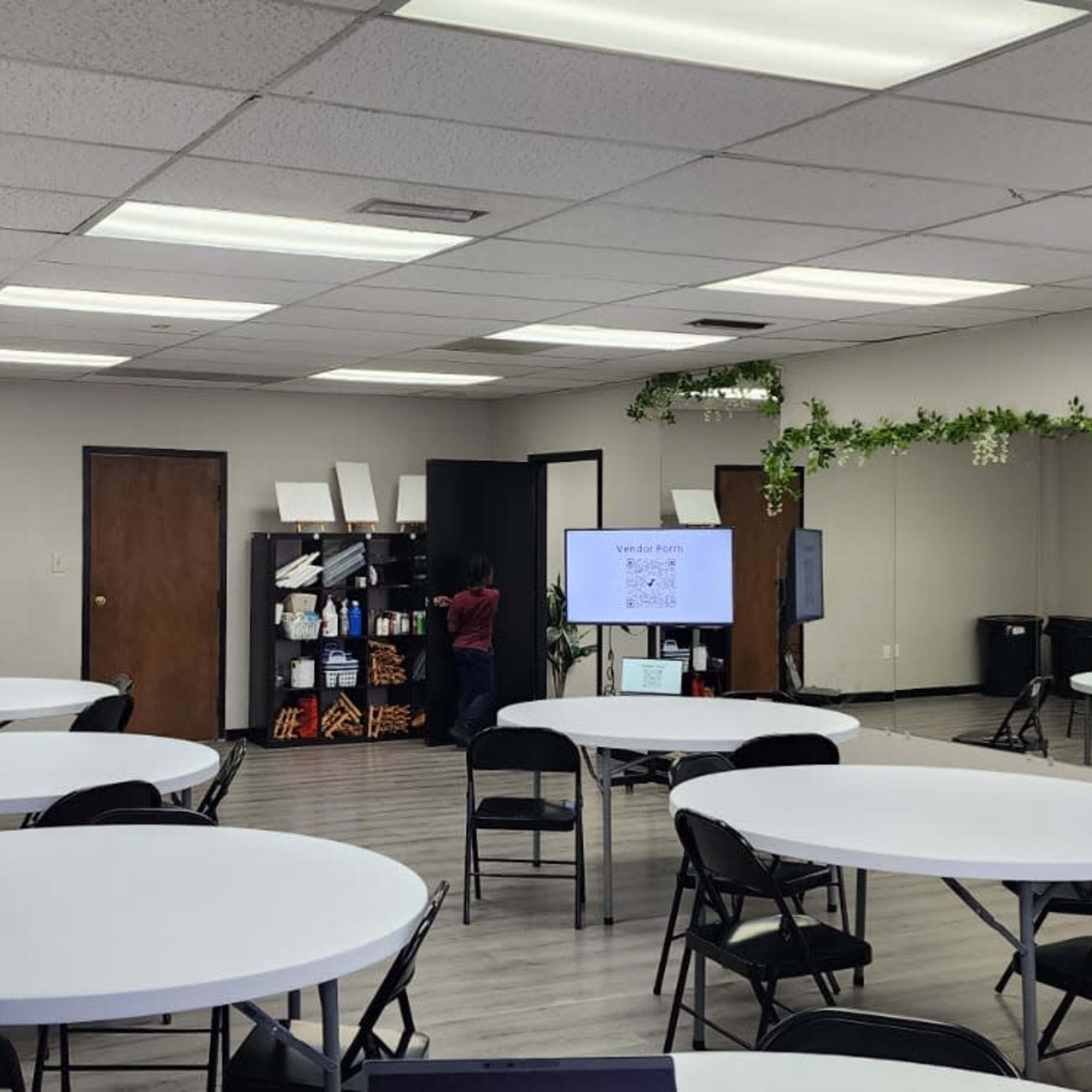 A spacious room with several round tables and chairs, a monitor displaying content, and a large mirror on one wall.
