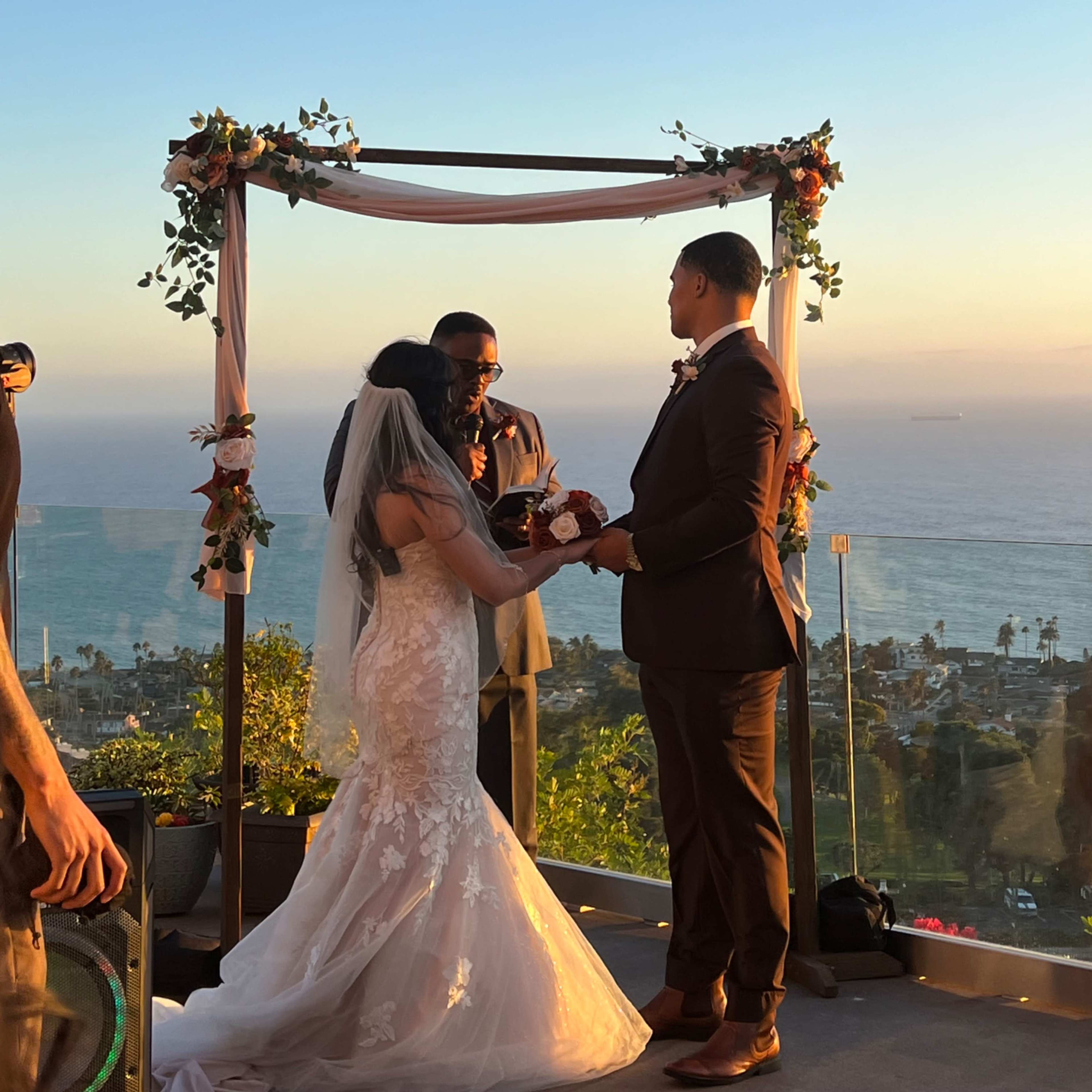 A couple exchanges vows during their wedding ceremony under an arch overlooking the ocean at sunset.