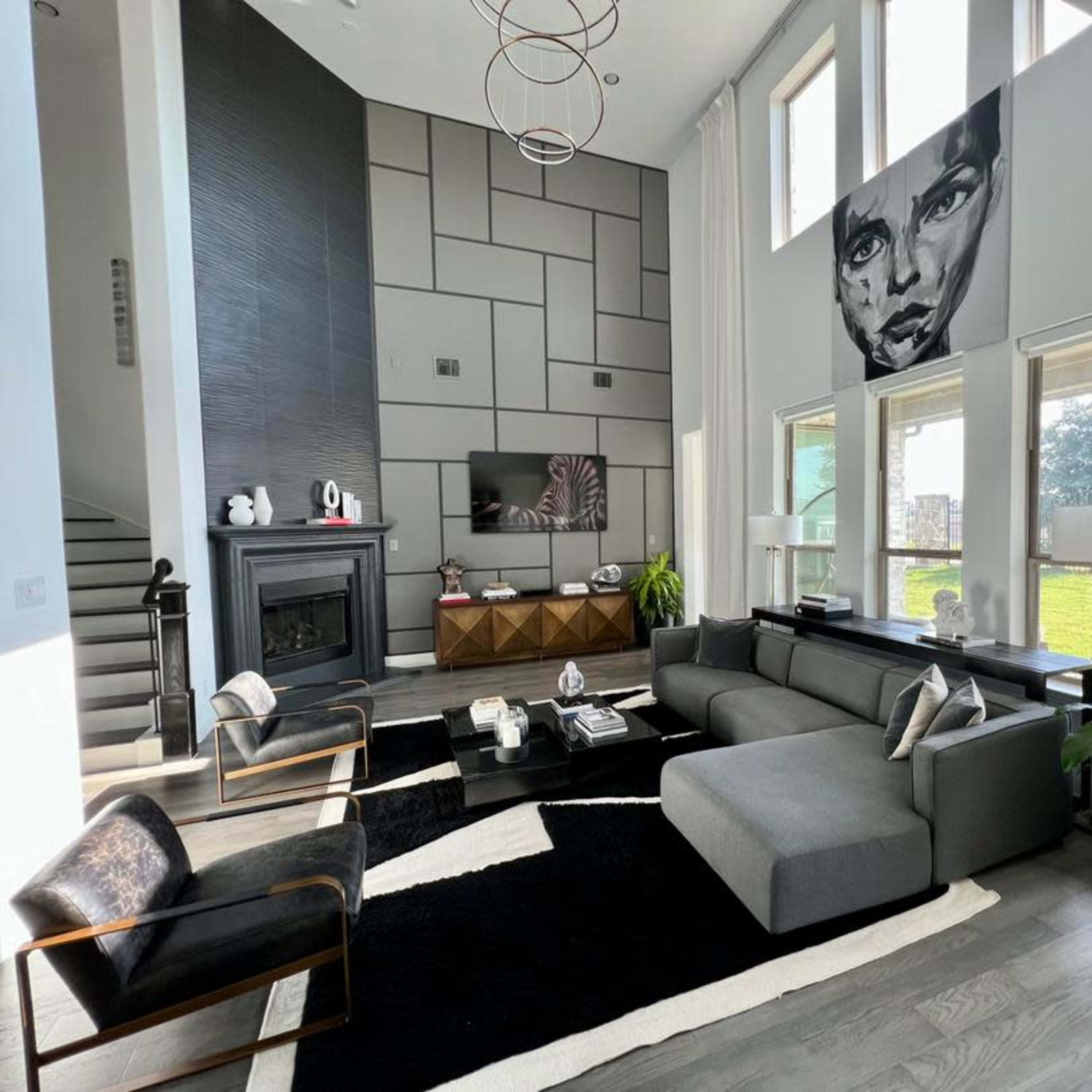 A modern living room with a large gray sectional sofa, black accent chairs, a coffee table, and a prominent wall art piece above a TV and fireplace, surrounded by large windows.
