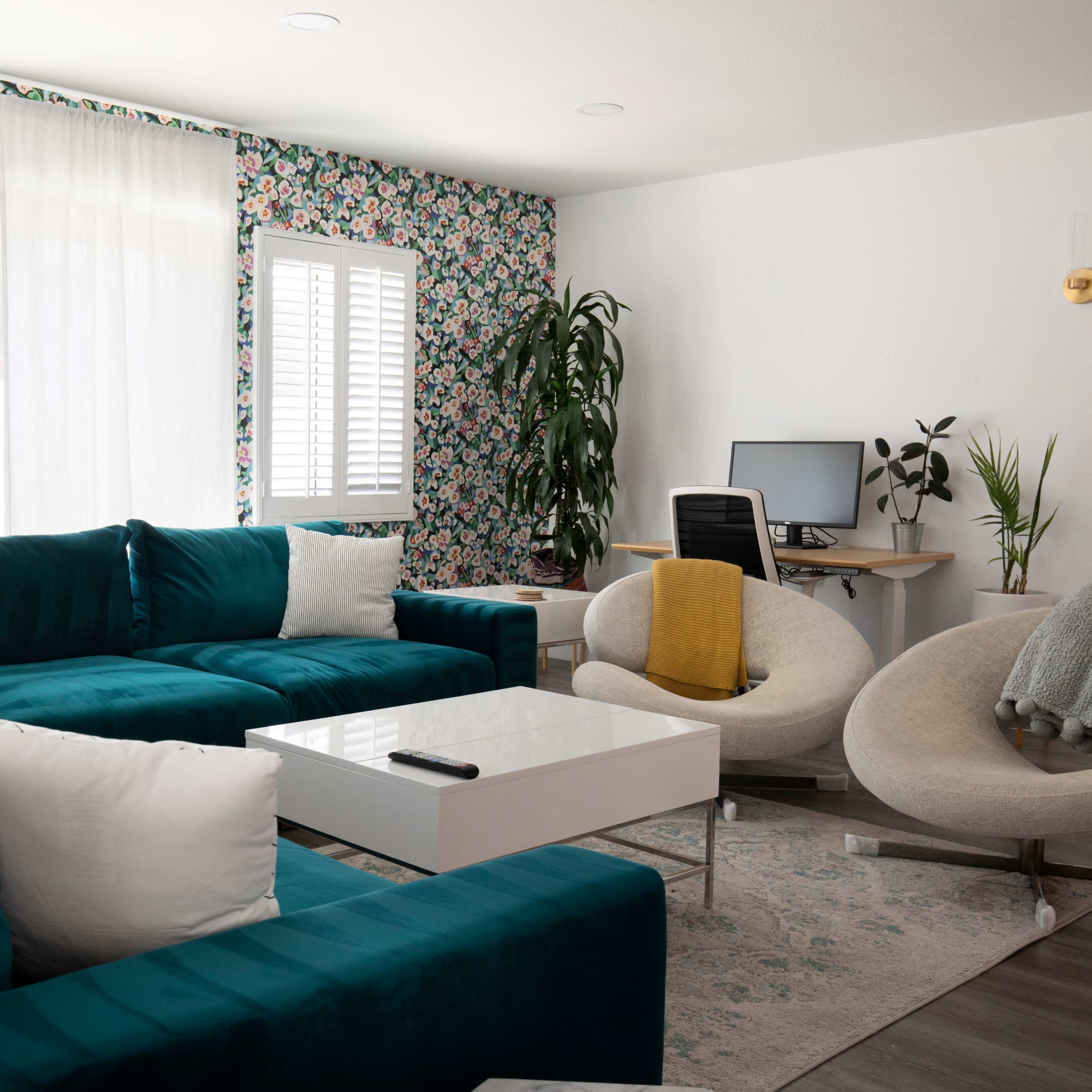 A modern living room with a teal sectional sofa, a white coffee table, a light-colored desk with a computer, and a decorative plant.