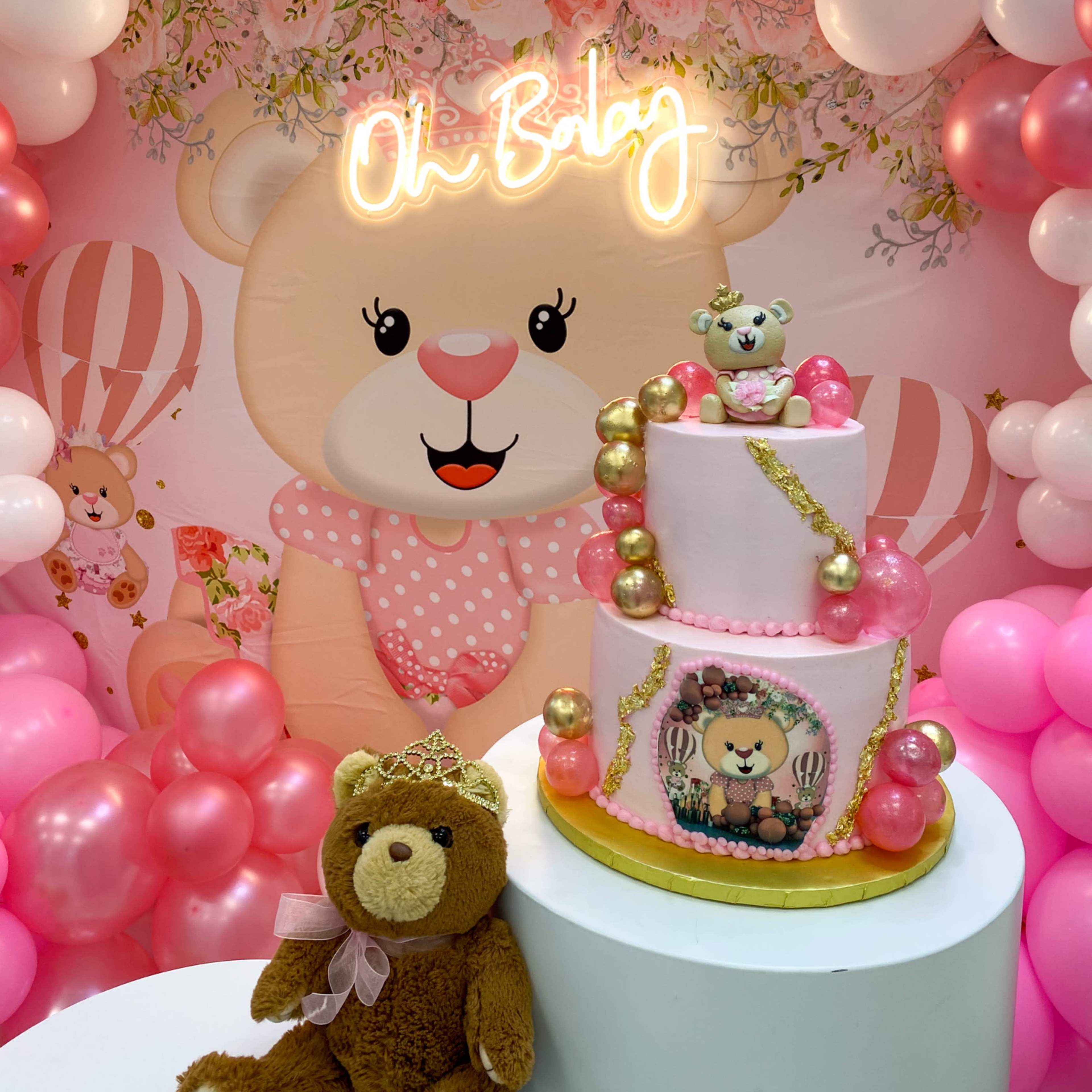 A three-tiered cake decorated with pink and gold accents is displayed in front of a balloon arch and a bear-themed backdrop with the words "Oh Baby."